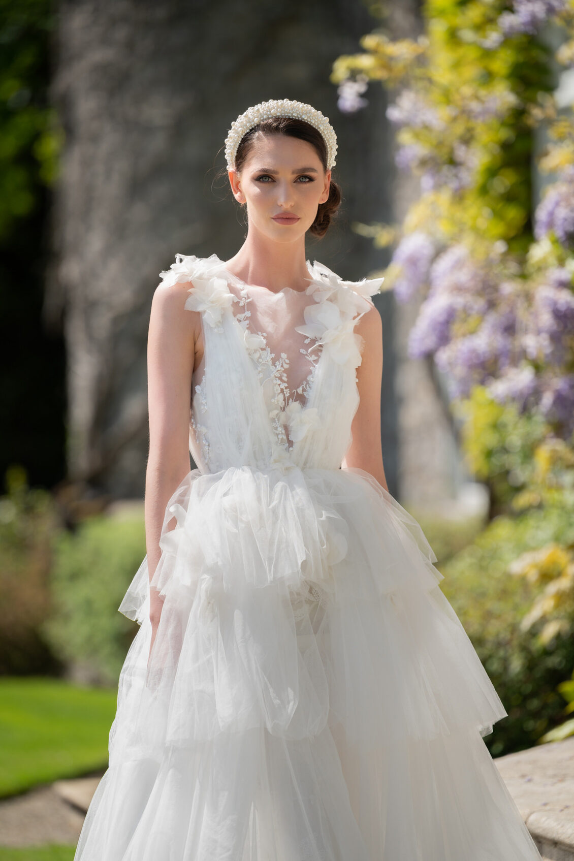 A bride in a sleeveless white wedding gown with a sheer floral bodice, layered tulle skirt, and pearl headband stands in a sunlit garden.