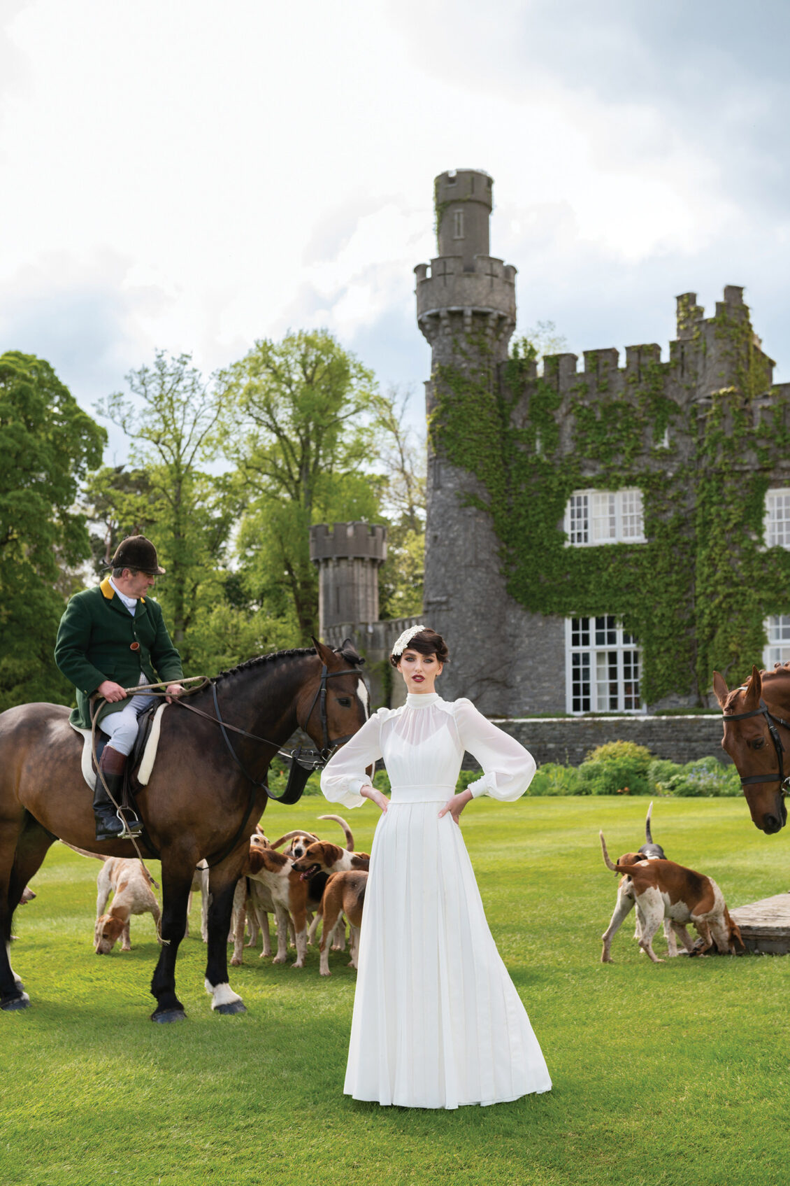 A bride in a white gown with sheer sleeves stands on a lawn beside a mounted rider, hounds, horses, and the ivy-covered Luttrellstown Castle.