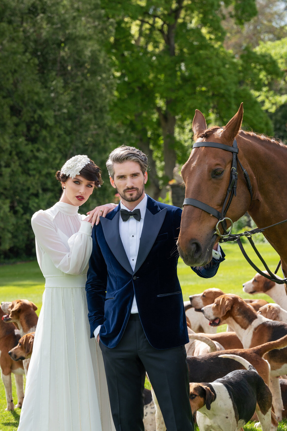 A bride in a long-sleeved white gown and beaded headpiece poses with a groom in a navy velvet tuxedo beside a brown horse and hounds.