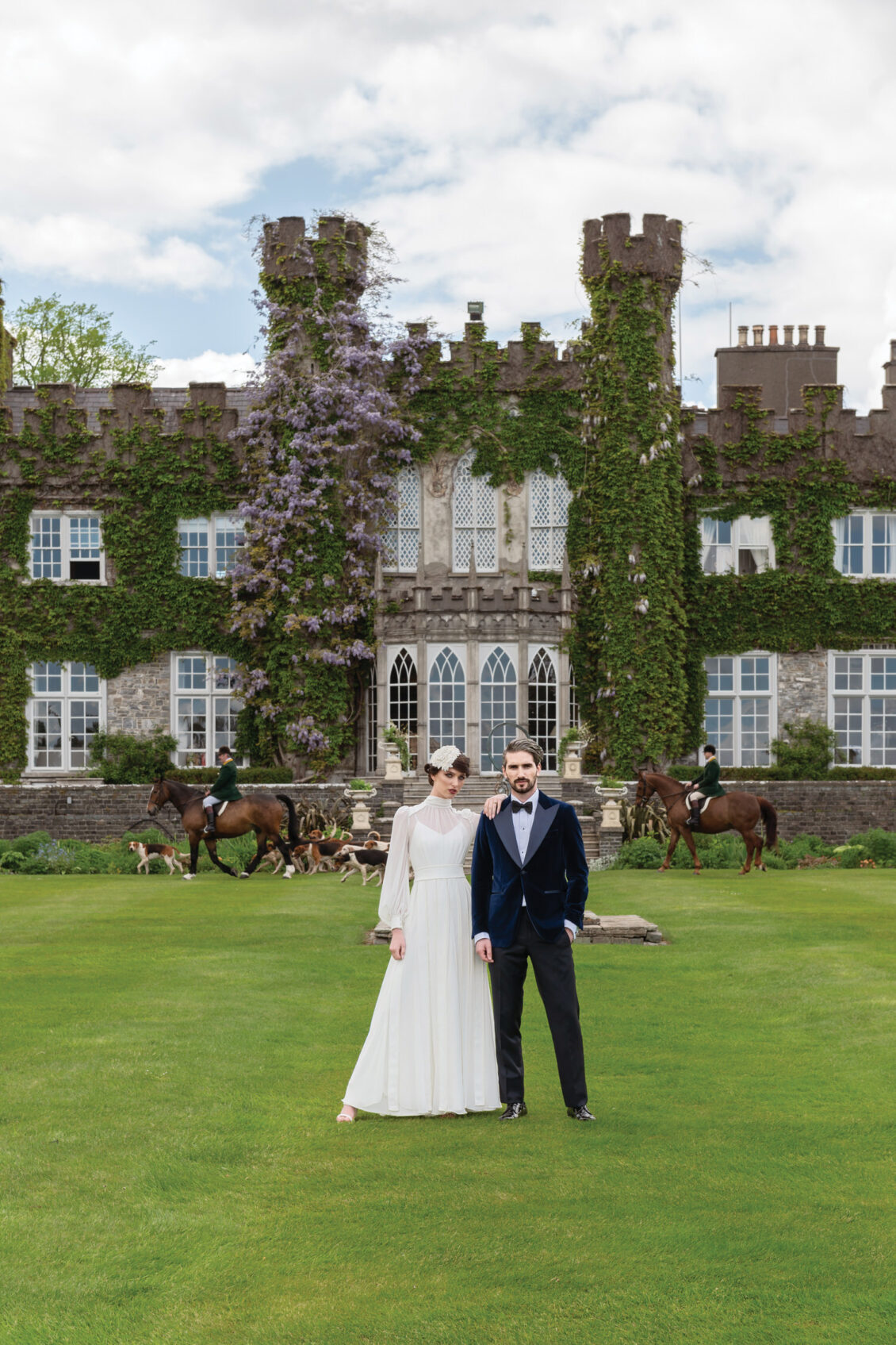 A bride and groom pose before the ivy-covered Luttrellstown Castle while two mounted riders flank a pack of hounds behind them.