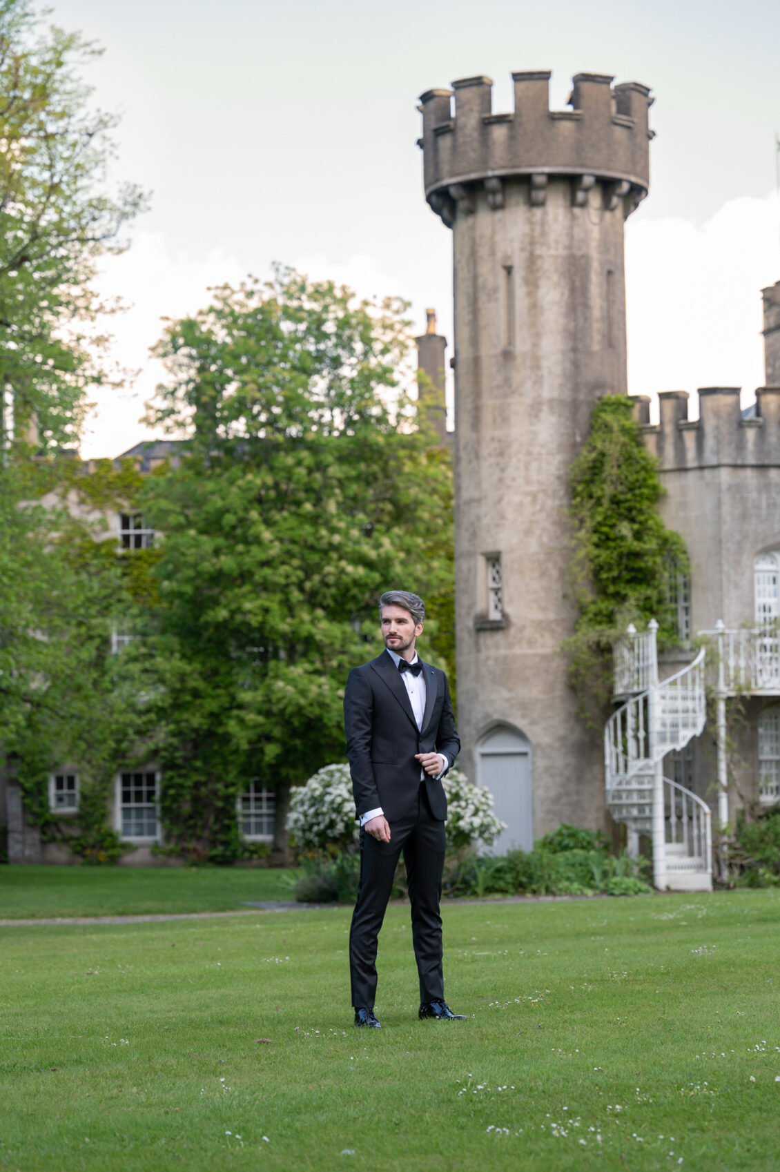 A groom in a black tuxedo and bow tie stands on the lawn before a stone castle with a round crenellated tower, ivy, and a white spiral staircase.