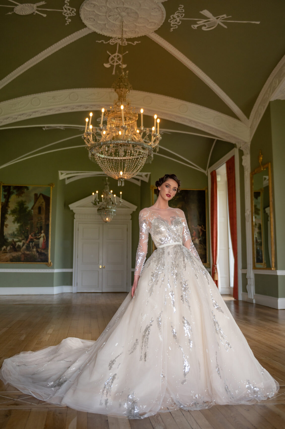 A bride in an ivory ball gown with silver floral embellishments stands beneath crystal chandeliers in an ornate green reception room.
