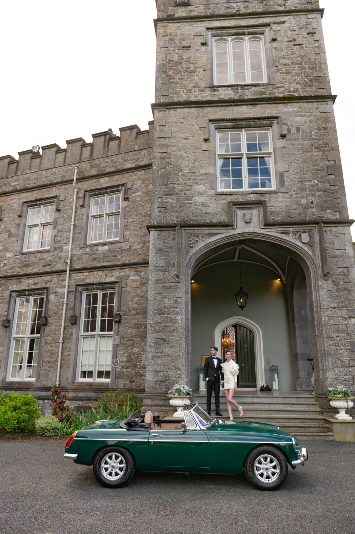 A couple holds hands on the steps of a stone manor beside a dark green vintage convertible; one wears a black tuxedo and the other a short white dress.