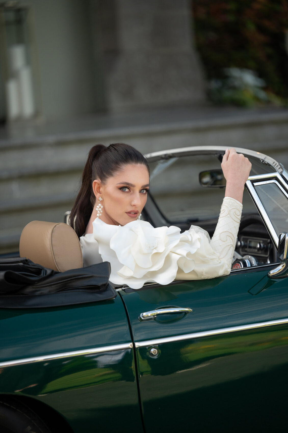 A bride in a long-sleeved white lace gown with oversized floral ruffles sits in a dark green vintage convertible outside a stone manor.