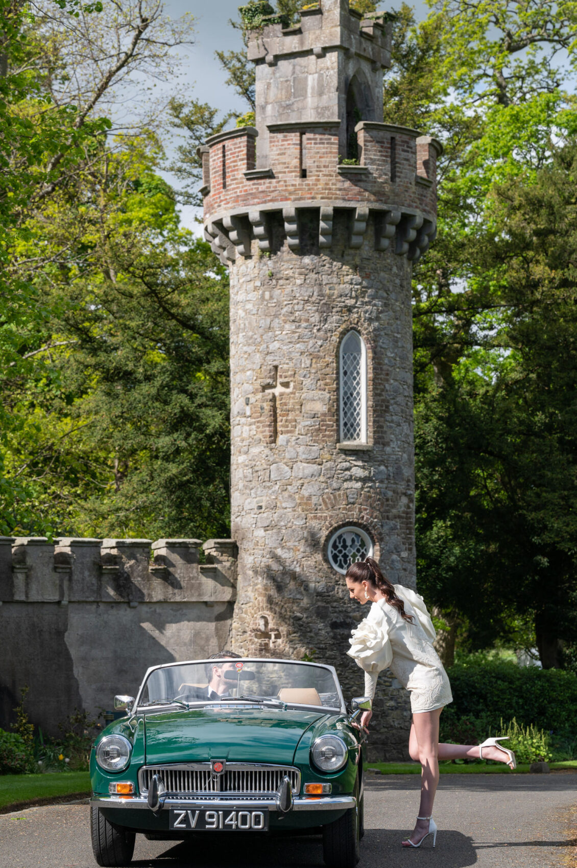 A bride in a short embroidered white dress leans into a vintage green convertible while a groom in a black tuxedo sits inside near a stone castle tower.