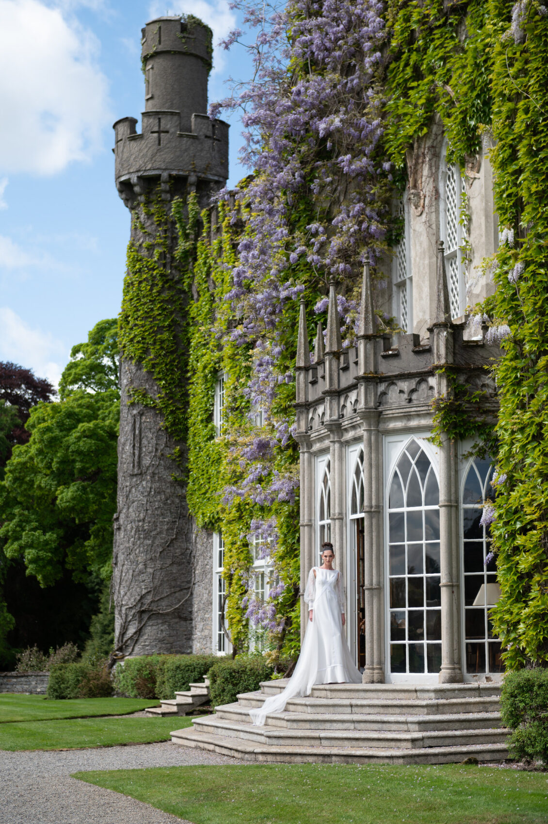 A woman in a long white wedding gown with sheer sleeves and a trailing train stands on stone steps outside an ivy-covered castle with arched windows and purple wisteria.