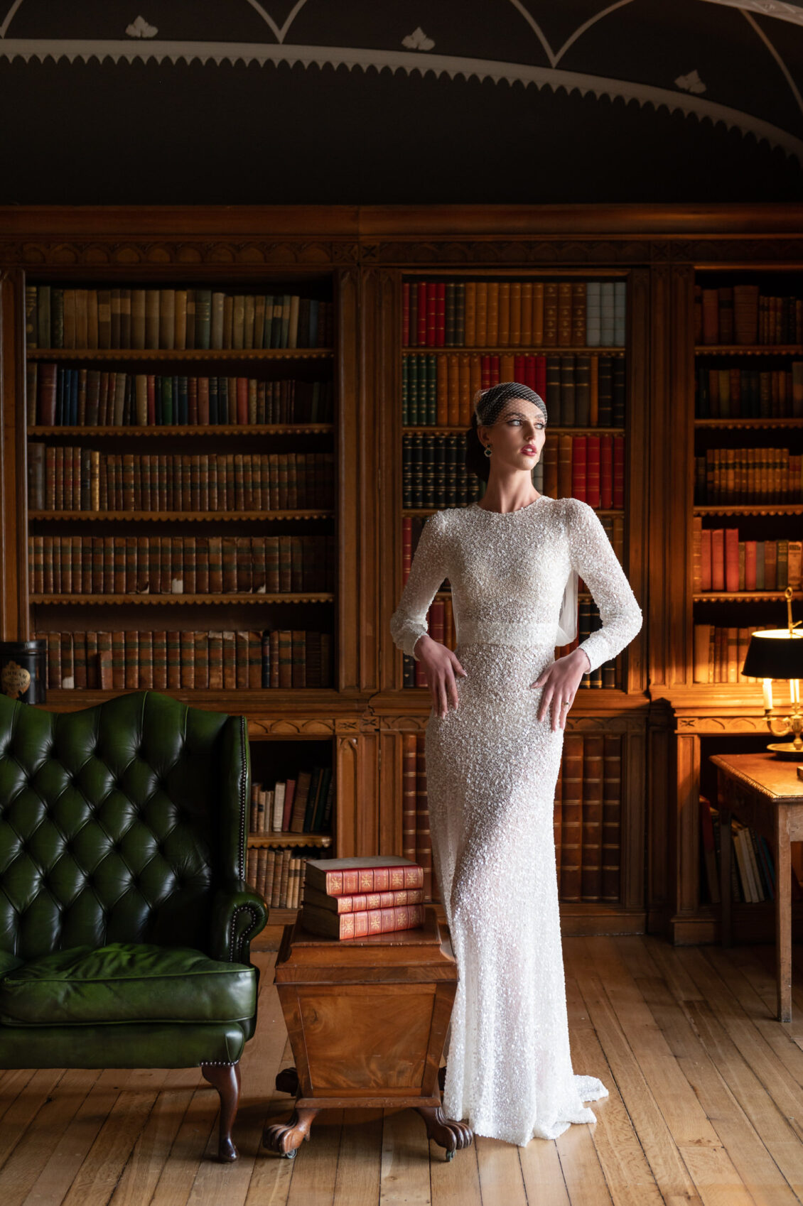 A bride in a fitted, long-sleeved white beaded gown and birdcage veil poses among floor-to-ceiling bookshelves in a wood-paneled library.