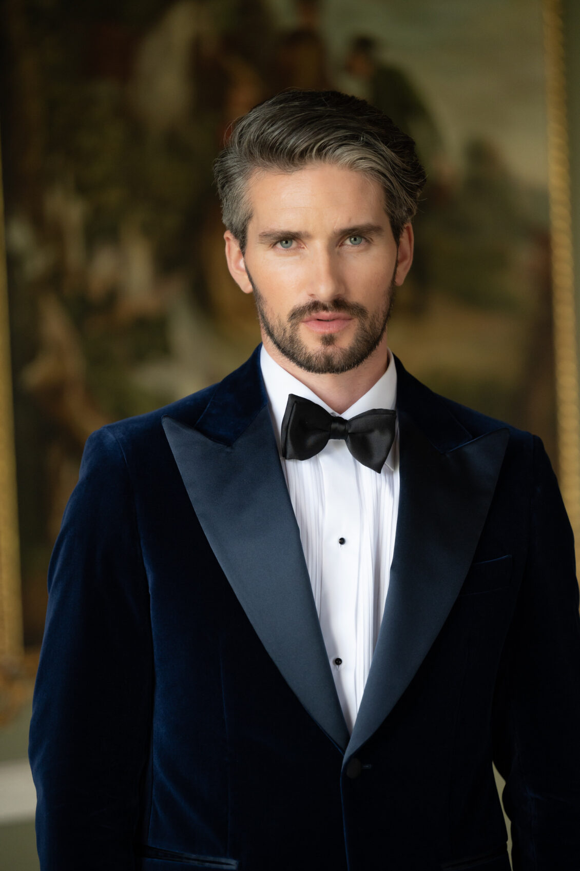 A bearded groom wearing a navy velvet tuxedo, white pleated shirt, and black bow tie poses indoors against a softly blurred gold-framed background.