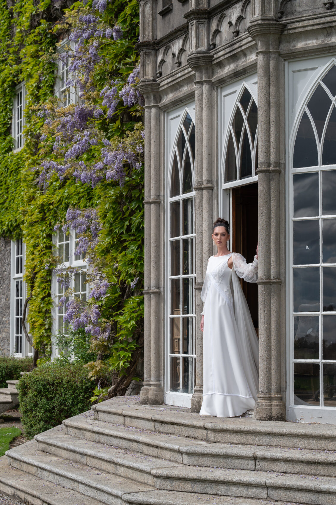 A woman in a floor-length white wedding gown with embroidered details and sheer sleeves stands in the open doorway of a stone manor surrounded by ivy and purple wisteria.