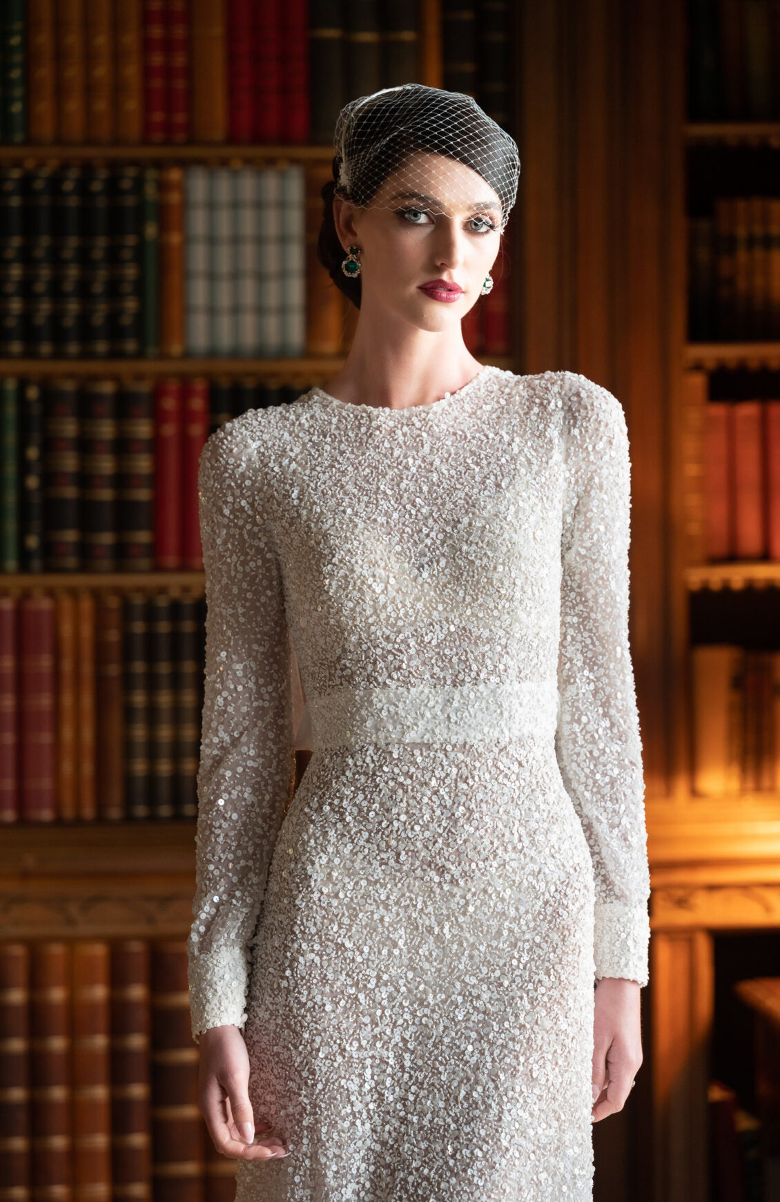 A woman in a long-sleeved, sequined white gown and a netted headpiece stands in front of bookshelves filled with books, softly lit by warm light—a scene reminiscent of the timeless elegance found at Luttrellstown Castle Resort’s Castle Keep, as featured in WedLuxe.