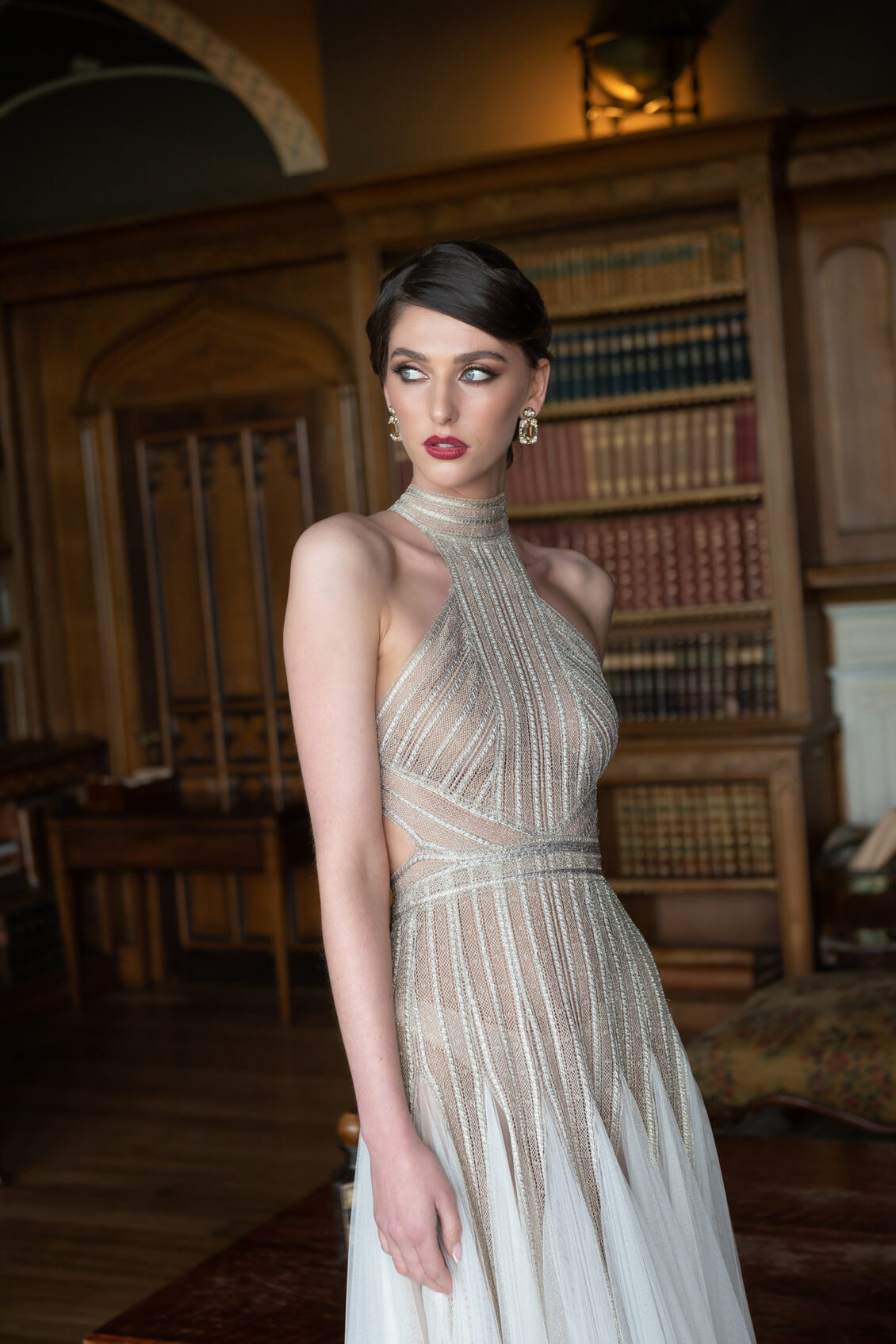 A bride in a high-neck wedding gown with metallic striped lace and a white tulle skirt stands beside wooden bookcases in a traditional library.
