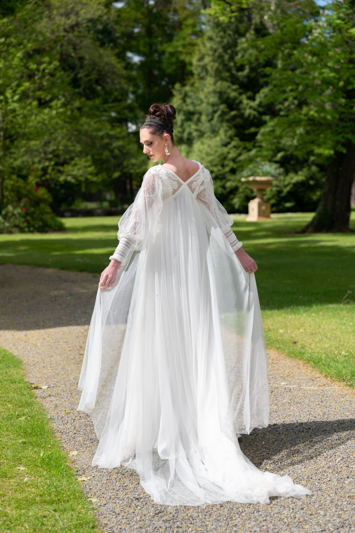 A bride in a flowing white wedding gown with sheer lace sleeves, an open V-back, and a long train stands on a gravel garden path.