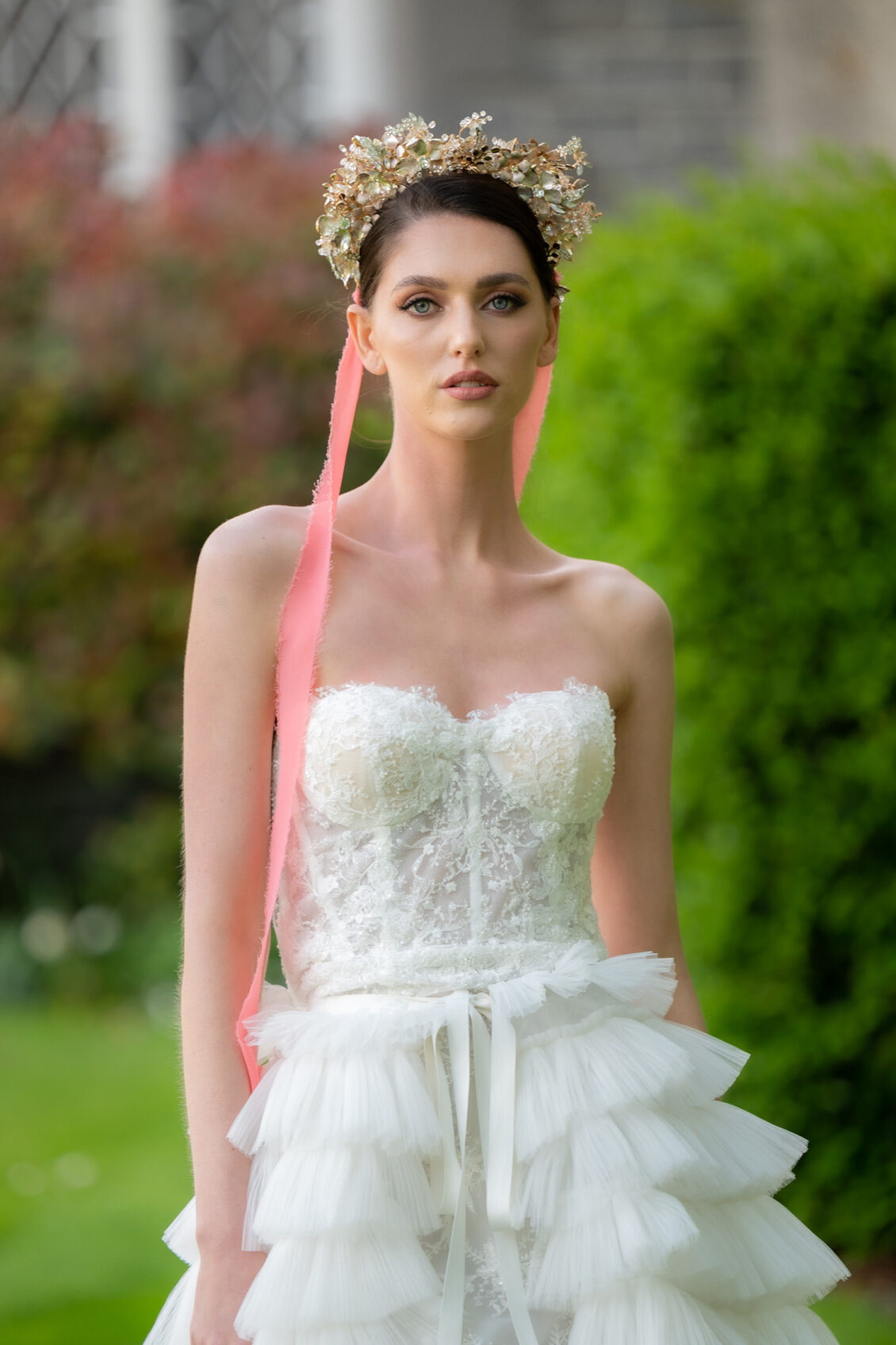 A bride in a strapless white lace corset gown and gold floral crown with long pink ribbons poses in a green garden.
