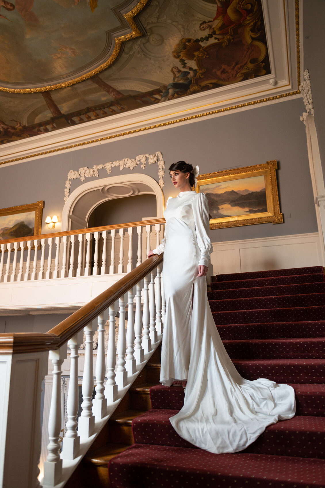 A bride in a long-sleeved white satin gown stands on a red-carpeted staircase with her train spread across the steps beneath a painted ceiling.