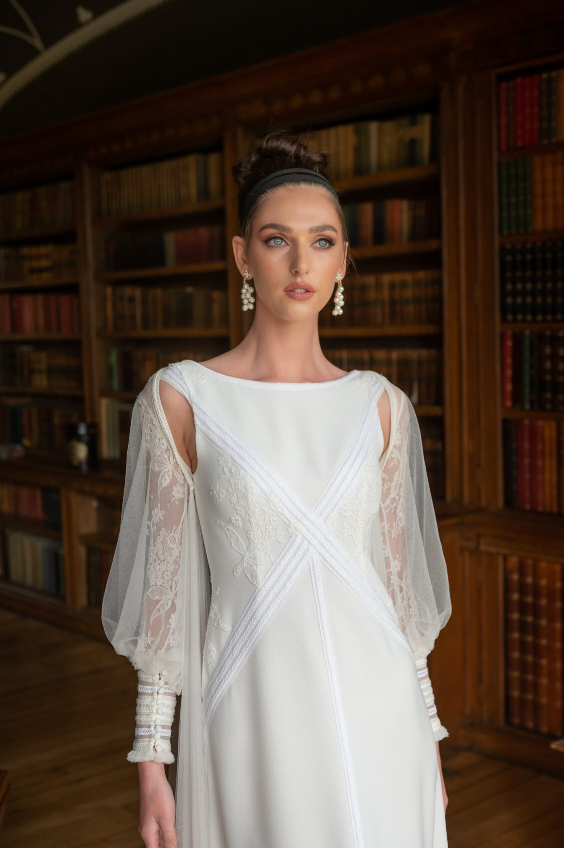 A bride in a floral-embroidered white wedding gown with sheer lace sleeves poses before book-filled shelves in a wood-paneled library.