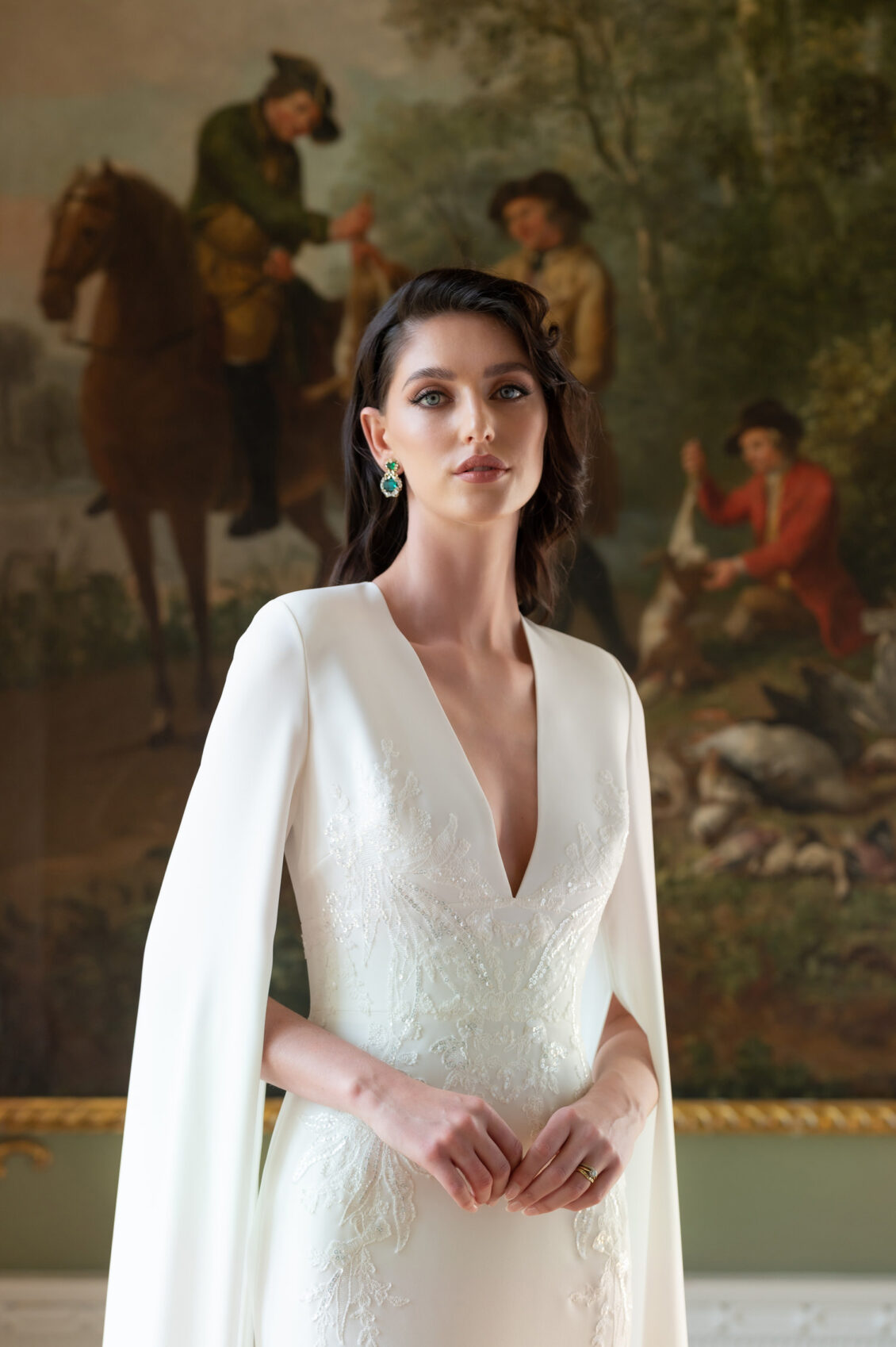 A bride wearing emerald earrings and a white deep-V wedding gown with lace embroidery and cape sleeves stands before a large equestrian painting.