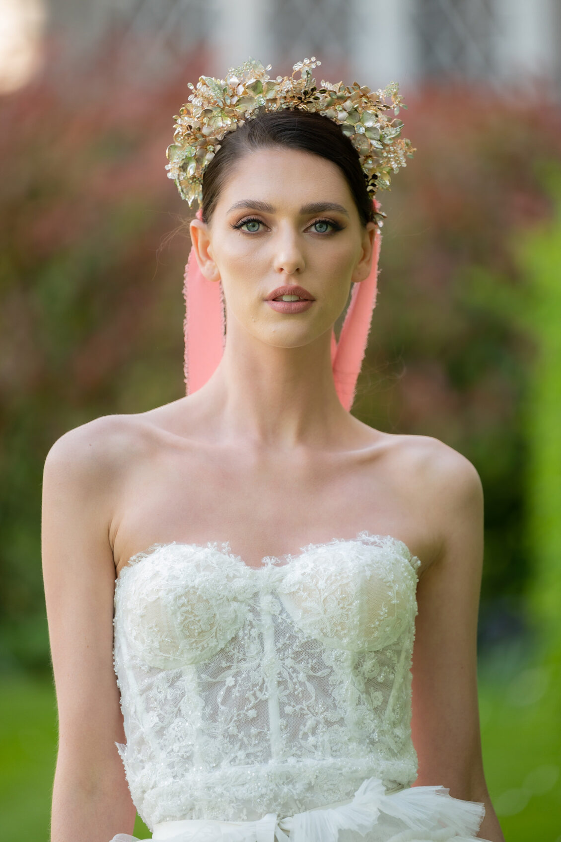 A bride in a strapless white lace wedding gown and ornate gold-and-green floral crown poses outdoors against softly blurred greenery.