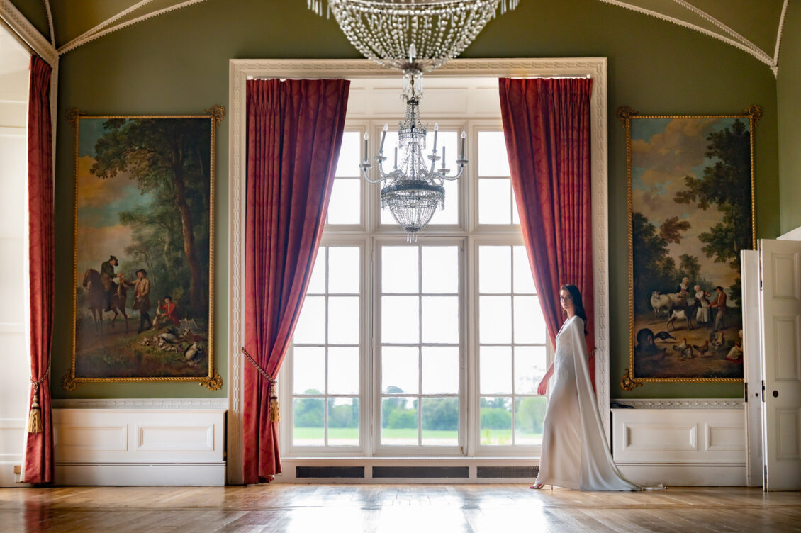 A woman in a white dress stands near tall windows with red curtains in an elegant room at Luttrellstown Castle Resort, featuring chandeliers and large classical paintings on the walls—a scene fit for the pages of WedLuxe.