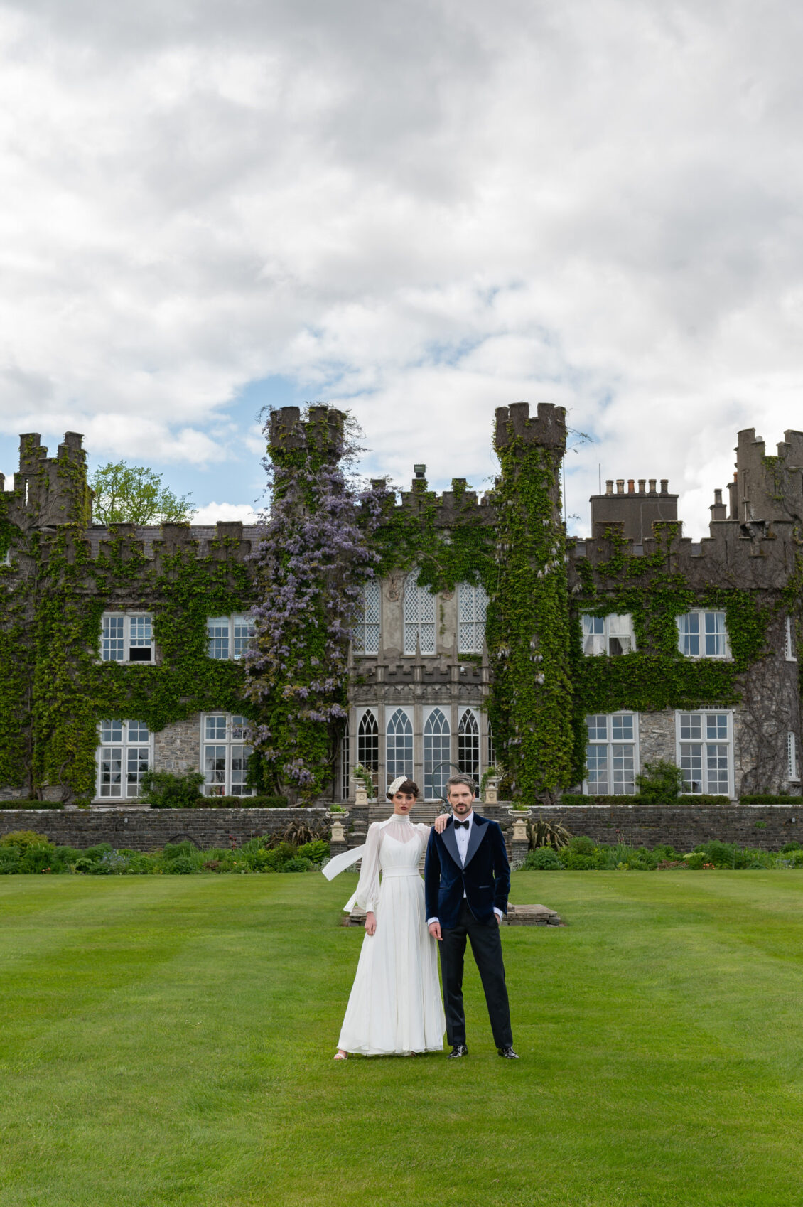 A bride in a long-sleeved white gown and floral headpiece rests her arm on a groom in a navy velvet tuxedo on the lawn at Luttrellstown Castle.