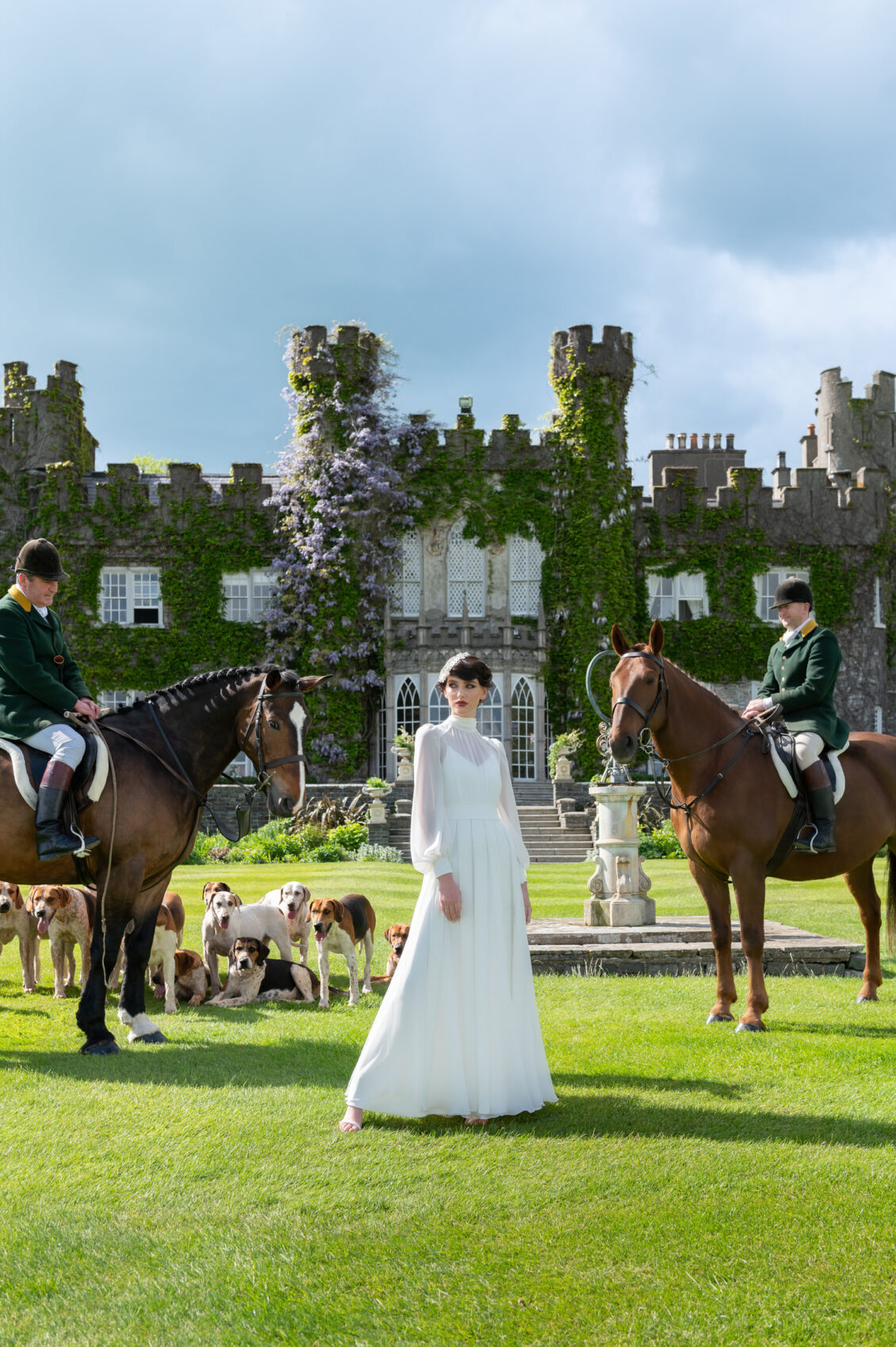 A woman in a white dress stands on grass before the enchanting Luttrellstown Castle Resort, flanked by two people on horses. Several dogs are gathered behind her, near a stone fountain, with the castle keep covered in ivy—a scene fit for a WedLuxe fairytale.