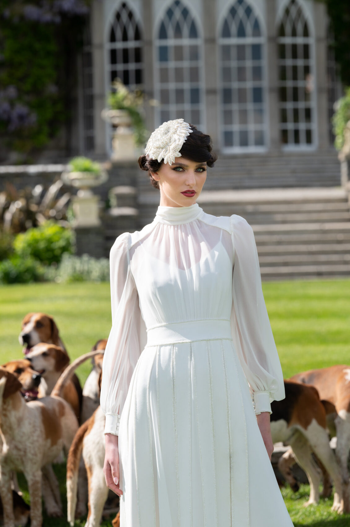 A woman in a vintage white dress and headpiece stands on a lawn in front of the grand Castle Keep at Luttrellstown Castle Resort, with a group of hounds elegantly assembled behind her—a scene reminiscent of WedLuxe-inspired romance.