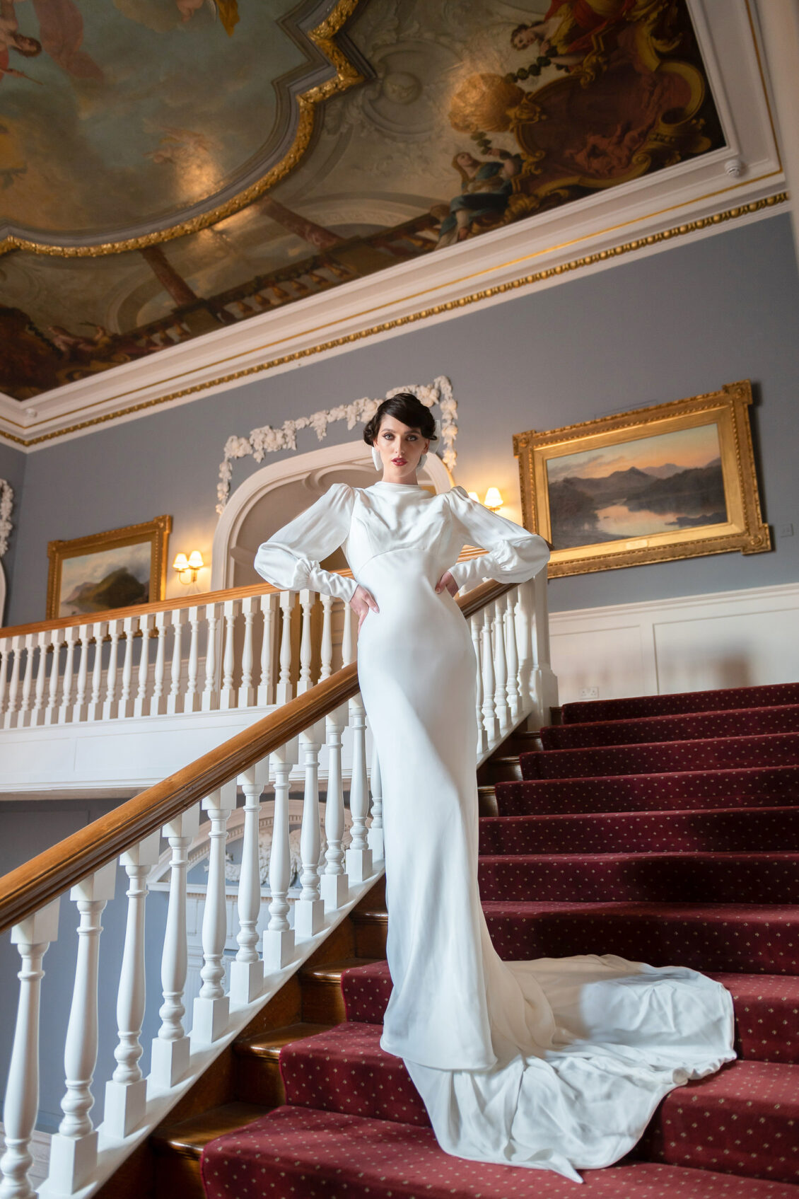 A woman wearing a long white gown stands on a grand staircase with red carpet, ornate railing, elegant paintings, and a decorative ceiling in the background, evoking the timeless romance of Luttrellstown Castle Resort.
