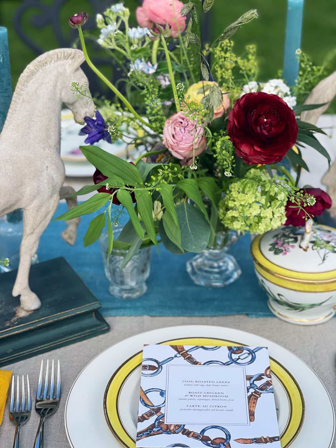 A close-up of an elegant table setting at Luttrellstown Castle, featuring a floral centerpiece, a white plate with a chain-patterned menu, silverware, a horse figurine on books, and a decorative lidded dish on a blue and beige tablecloth—perfect inspiration for your dream wedding venue.