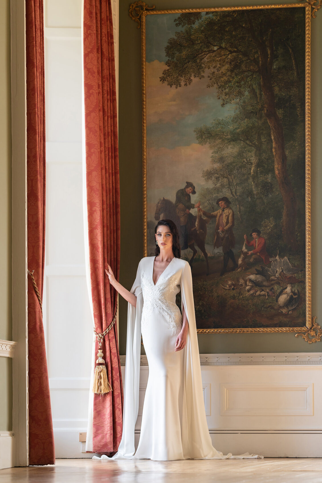 A woman in a white, elegant gown stands by red curtains in a grand room at Luttrellstown Castle, with a large, classical painting on the wall behind her—an exquisite setting that perfectly captures the timeless charm of this renowned wedding venue.