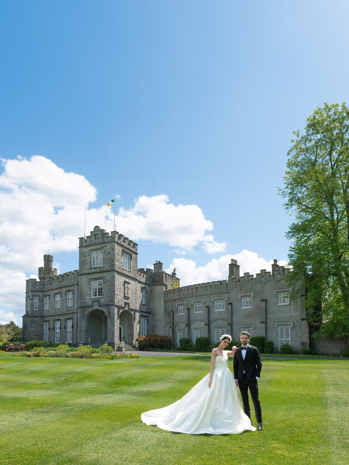A bride and groom stand on a green lawn in front of the majestic Luttrellstown Castle Resort, under a blue sky with scattered clouds.