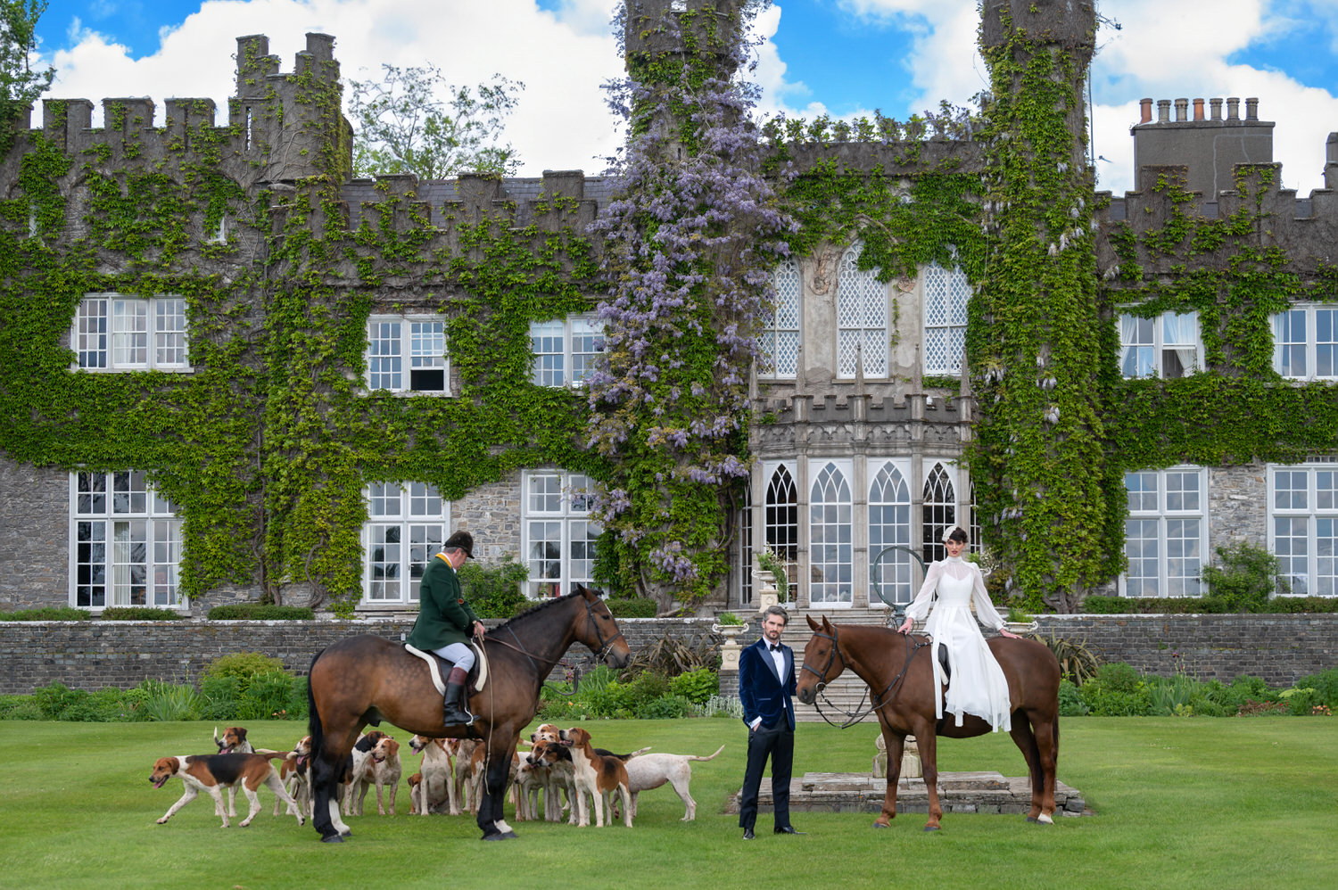 Two people on horses and a man standing between them pose in front of ivy-covered Luttrellstown Castle, with several dogs gathered around on the green lawn. The scene is formal, elegant, and outdoors—evoking the refined atmosphere of a castle wedding worthy of WedLuxe.