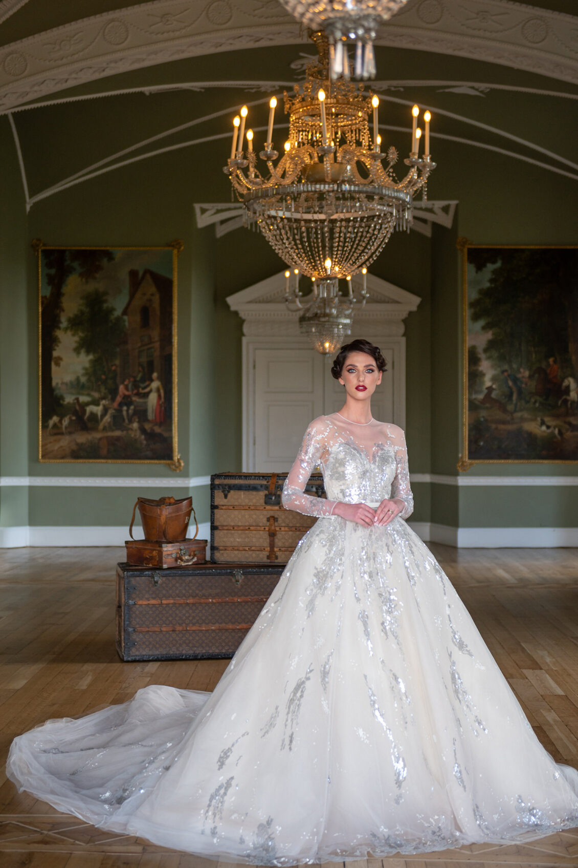 A woman in a white, sparkling ball gown stands in an elegant room with green walls, chandeliers, large paintings, and antique trunks at the renowned Luttrellstown Castle Resort. The enchanting setting exudes fairytale charm worthy of WedLuxe, perfectly blending timeless elegance with modern romance.