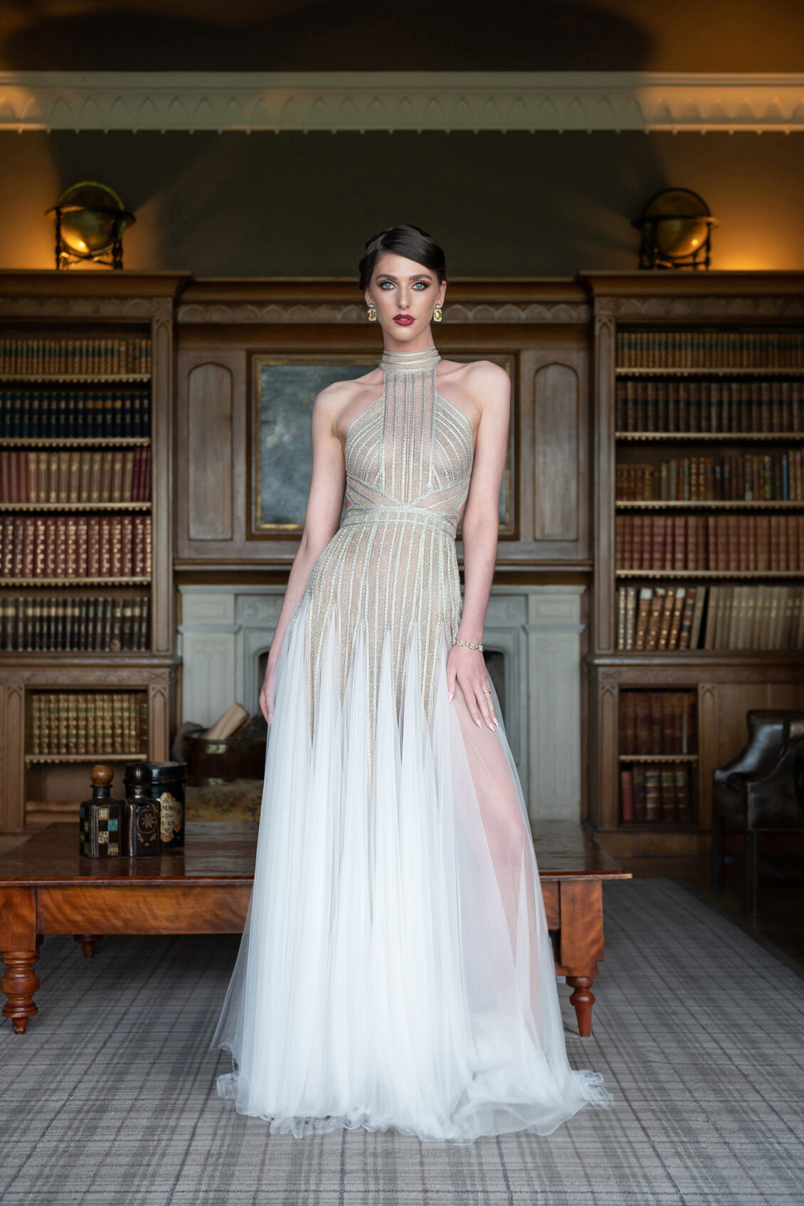 A woman in a sleeveless, white and gold evening gown stands in a library with wooden bookshelves, a wooden table, and soft lighting, evoking the timeless elegance of a Luttrellstown Castle Resort castle wedding as featured in WedLuxe.