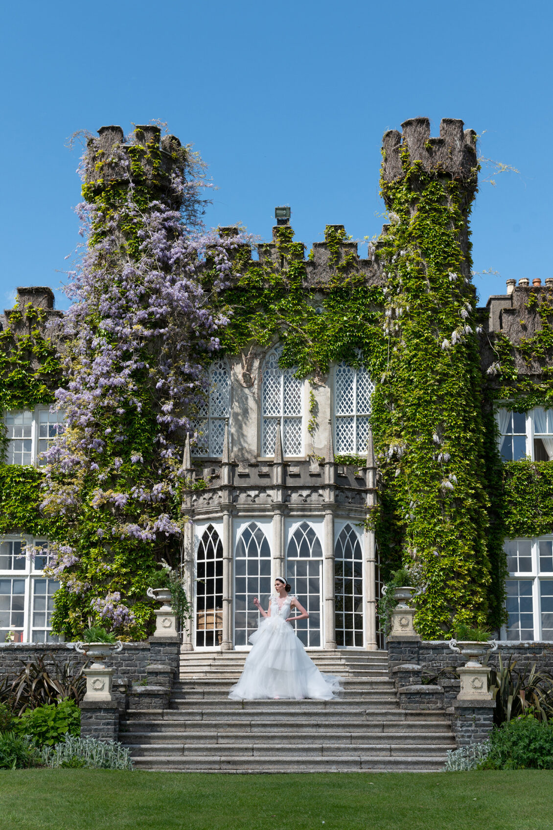 A bride in a white gown stands on stone steps in front of the grand Luttrellstown Castle keep, its tall towers and arched windows framed by lush ivy, all beneath a clear blue sky—a scene fit for the pages of WedLuxe.