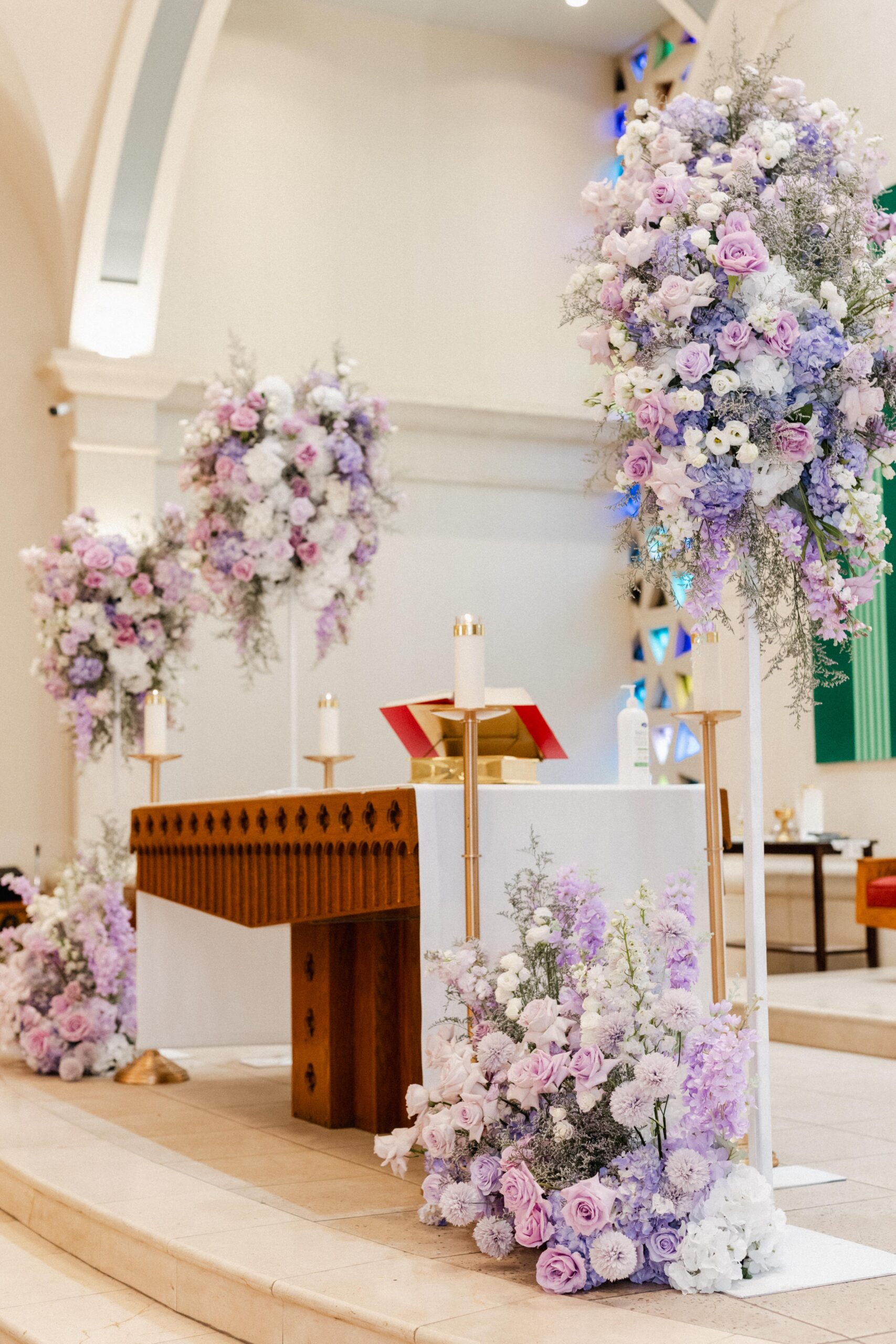 An ornate church altar adorned with tall floral arrangements featuring  white, purple, and lilac flowers. Candles, image size:1707x2560