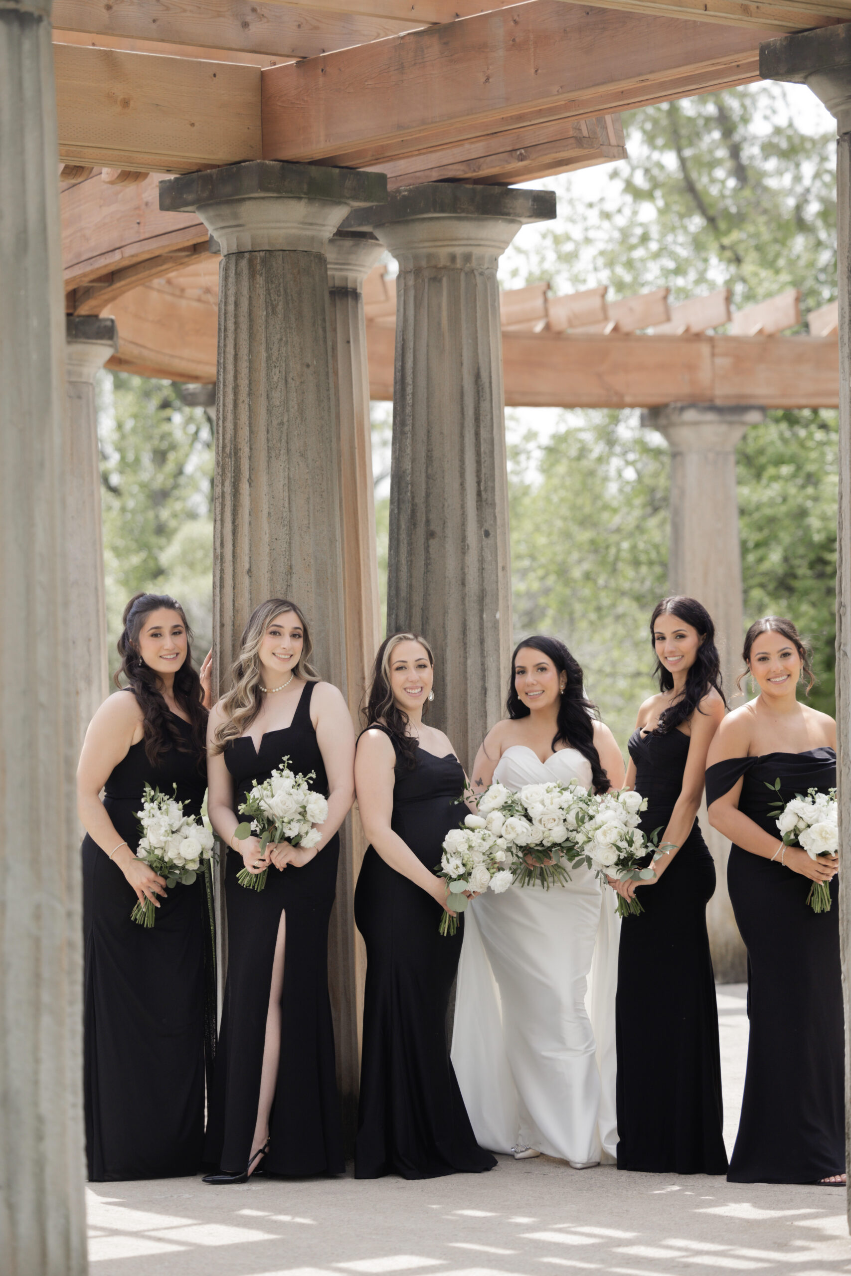 Six women stand under a pergola with columns around them. In this modern woodland  wedding setting,