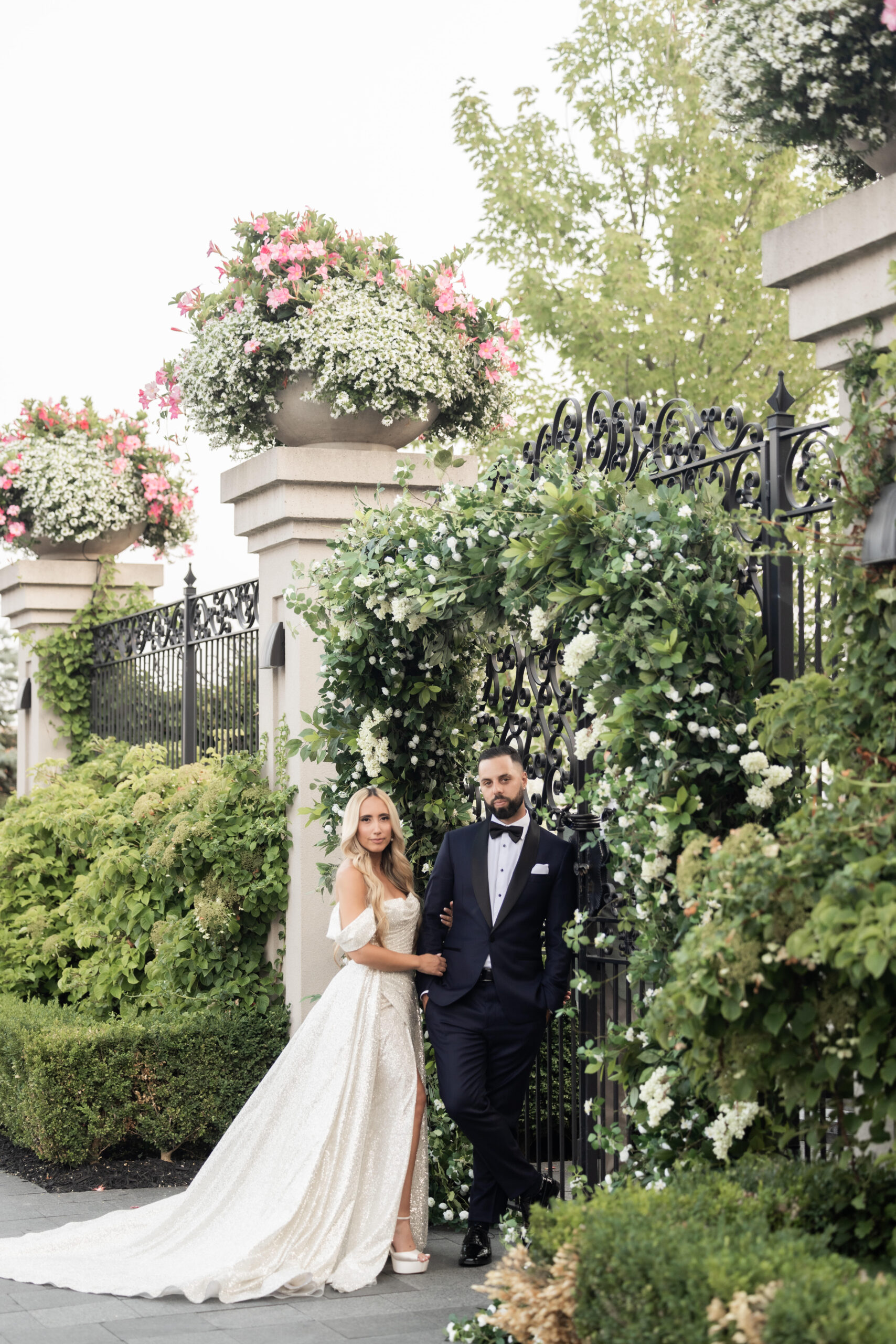 A couple in formal attire stands by an ornate wrought iron fence, lush with  greenery and