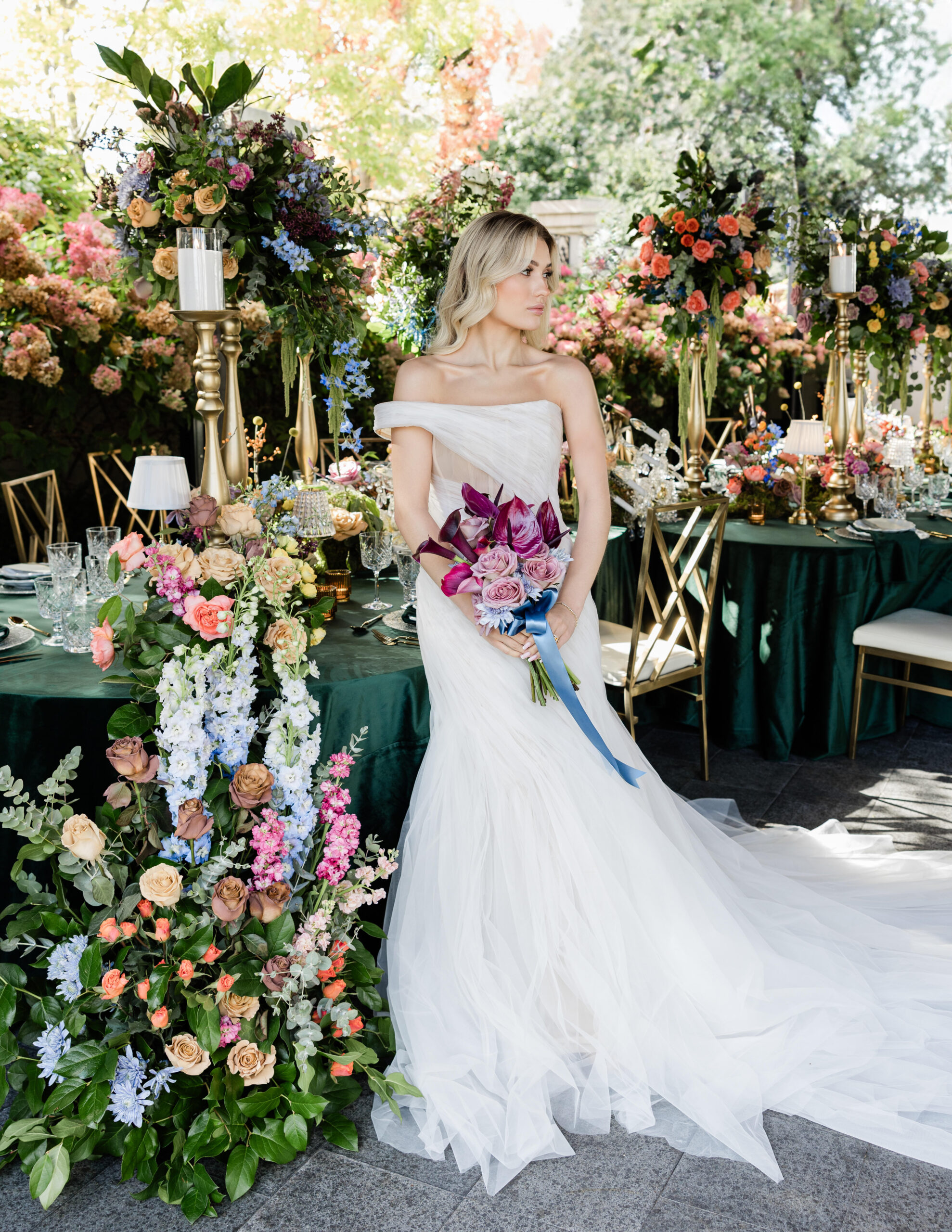 The bride, in an off-the-shoulder white gown, holds a bouquet with purple  flowers, standing