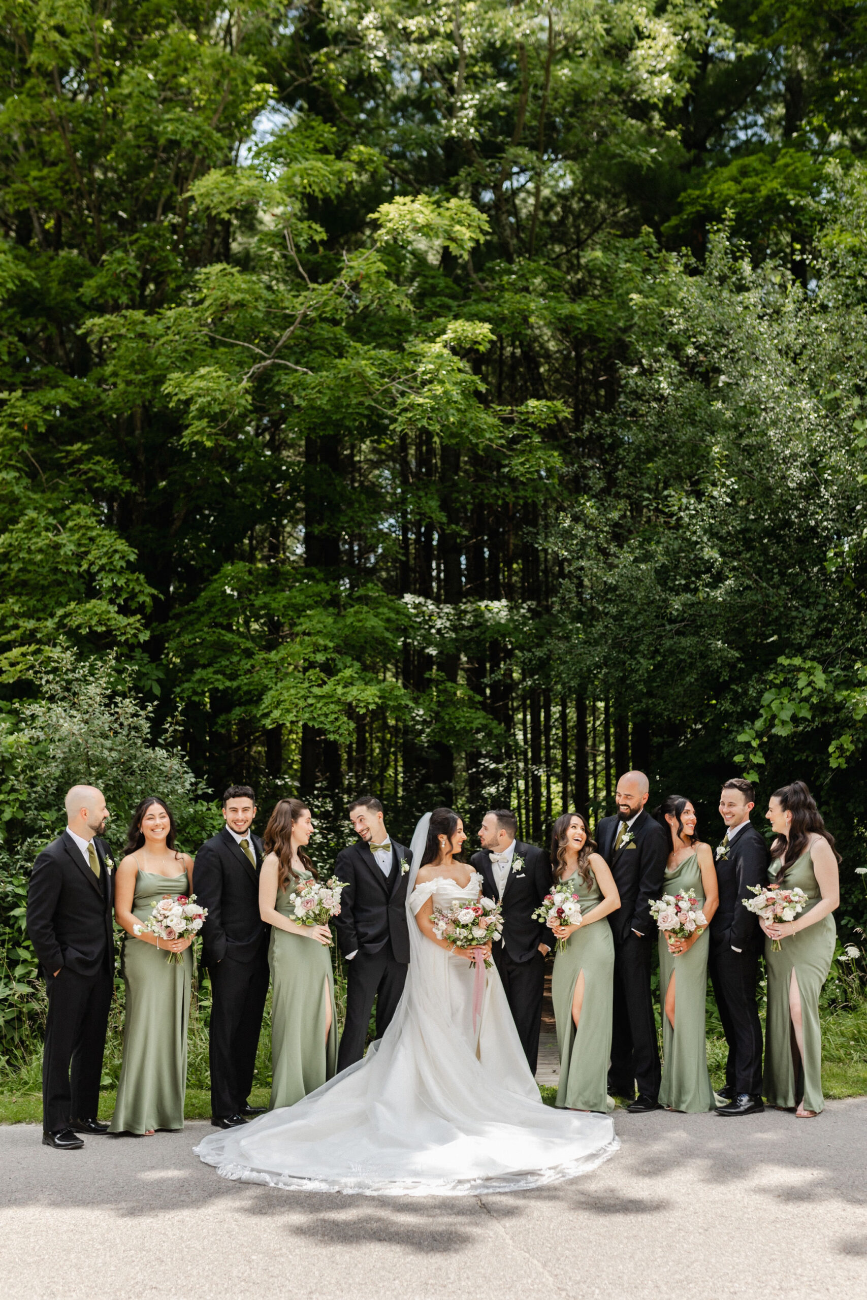 sage green bridesmaid dresses with black suits A wedding party stands outdoors, smiling and laughing at a European garden  wedding. The bride in