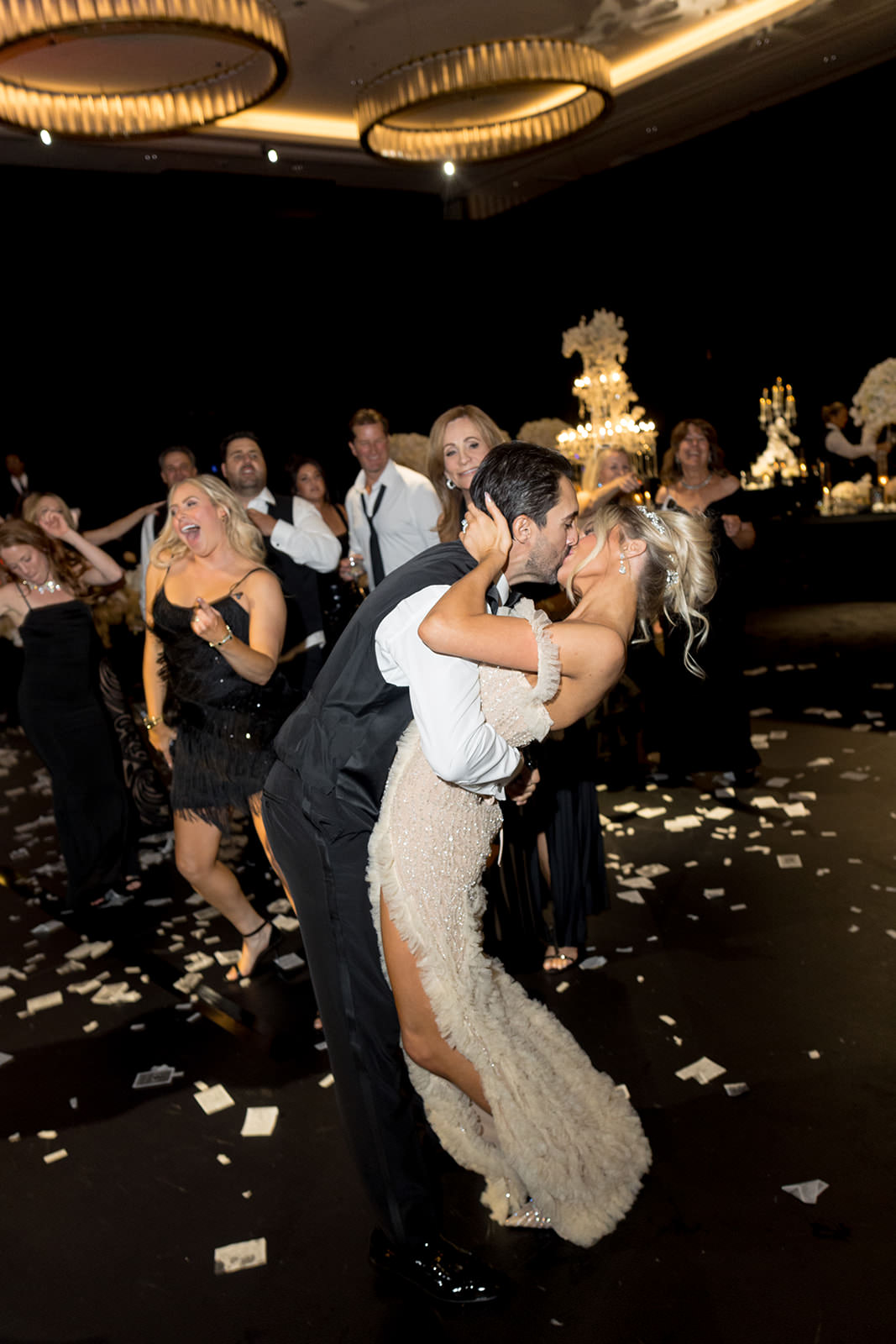 A bride and groom share a joyful kiss and dip on a confetti-covered dance floor at Fontainebleau Las Vegas, surrounded by cheering wedding guests in formal attire under elegant lighting. ~ WedLuxe A bride and groom share a joyful kiss and dip on a confetti-covered dance floor at Fontainebleau Las Vegas, surrounded by cheering wedding guests in formal attire under elegant lighting. ~ WedLuxe