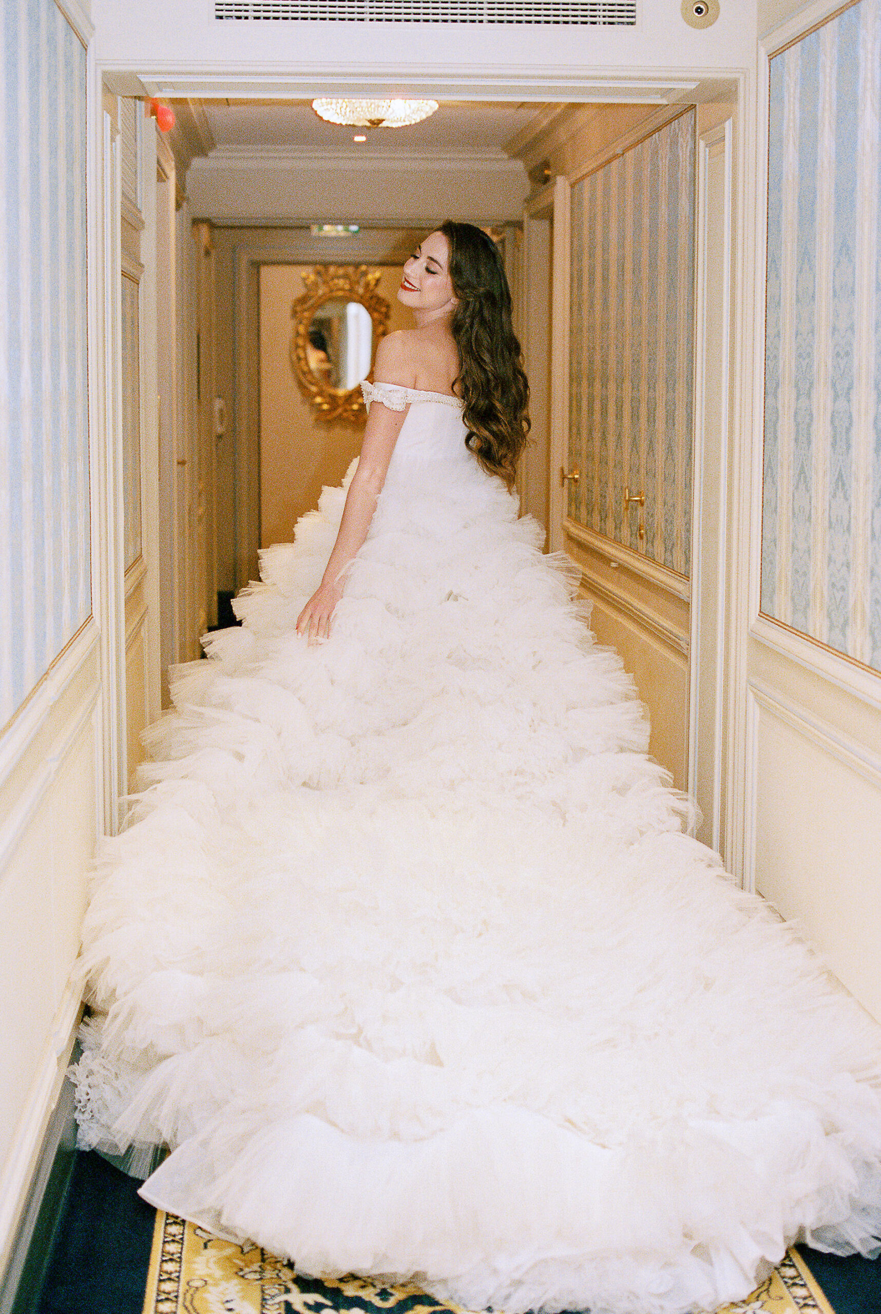 script src="https://form.jotform.com/jsform/261124848760157"  defer></script>A woman in an off-the-shoulder, voluminous white wedding gown  with a long ruffled train stands in an elegant hallway at The Ritz, Paris,  looking back over her shoulder and smiling.