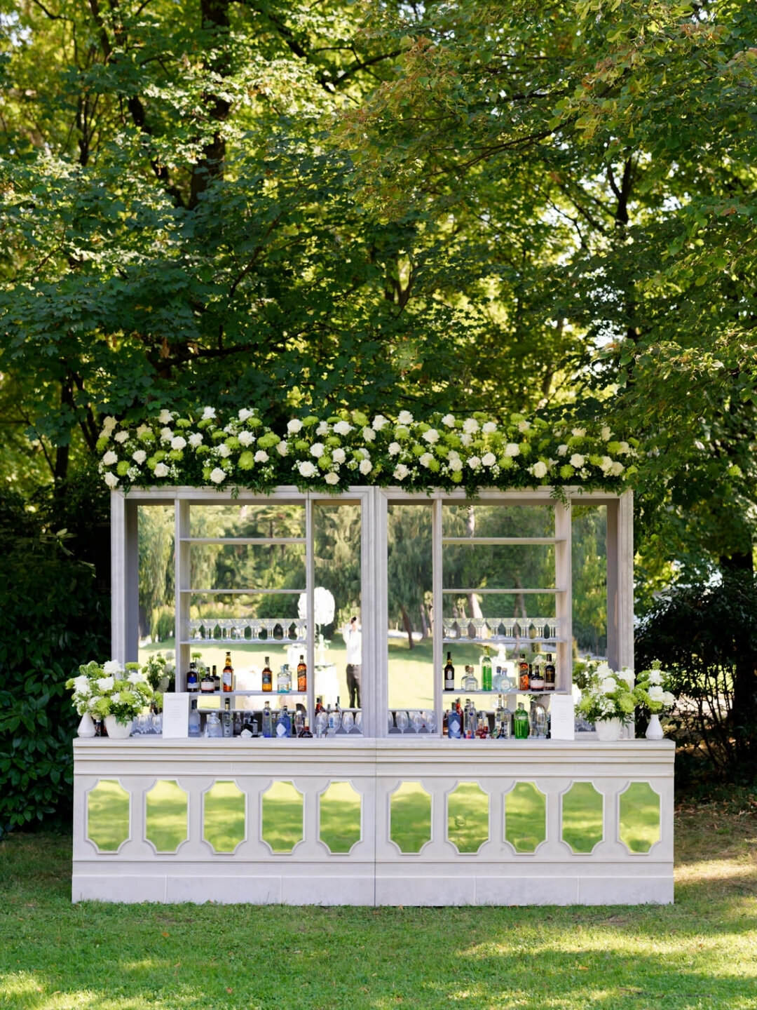 A stylish outdoor bar at Villa Erba, decorated with white flowers and greenery, stocked with bottles and glassware, set against the lush green backdrop of Lake Como—perfect for an elegant Italian wedding.