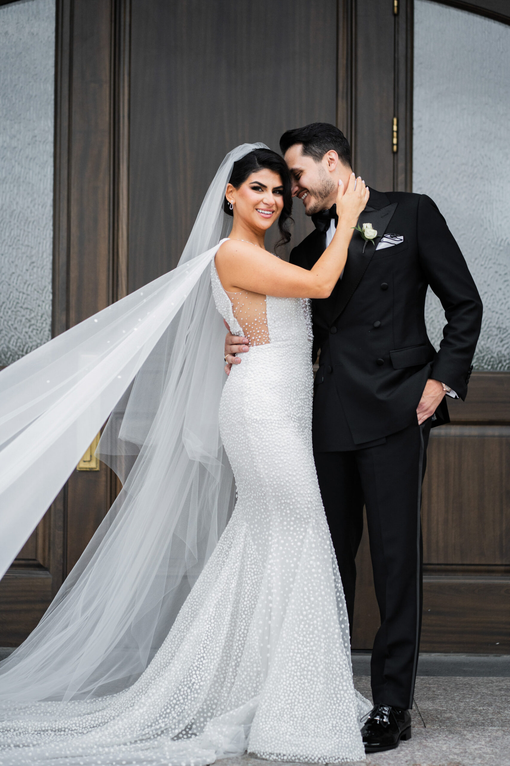 A bride in a fitted, beaded white gown and long veil stands smiling next to  a