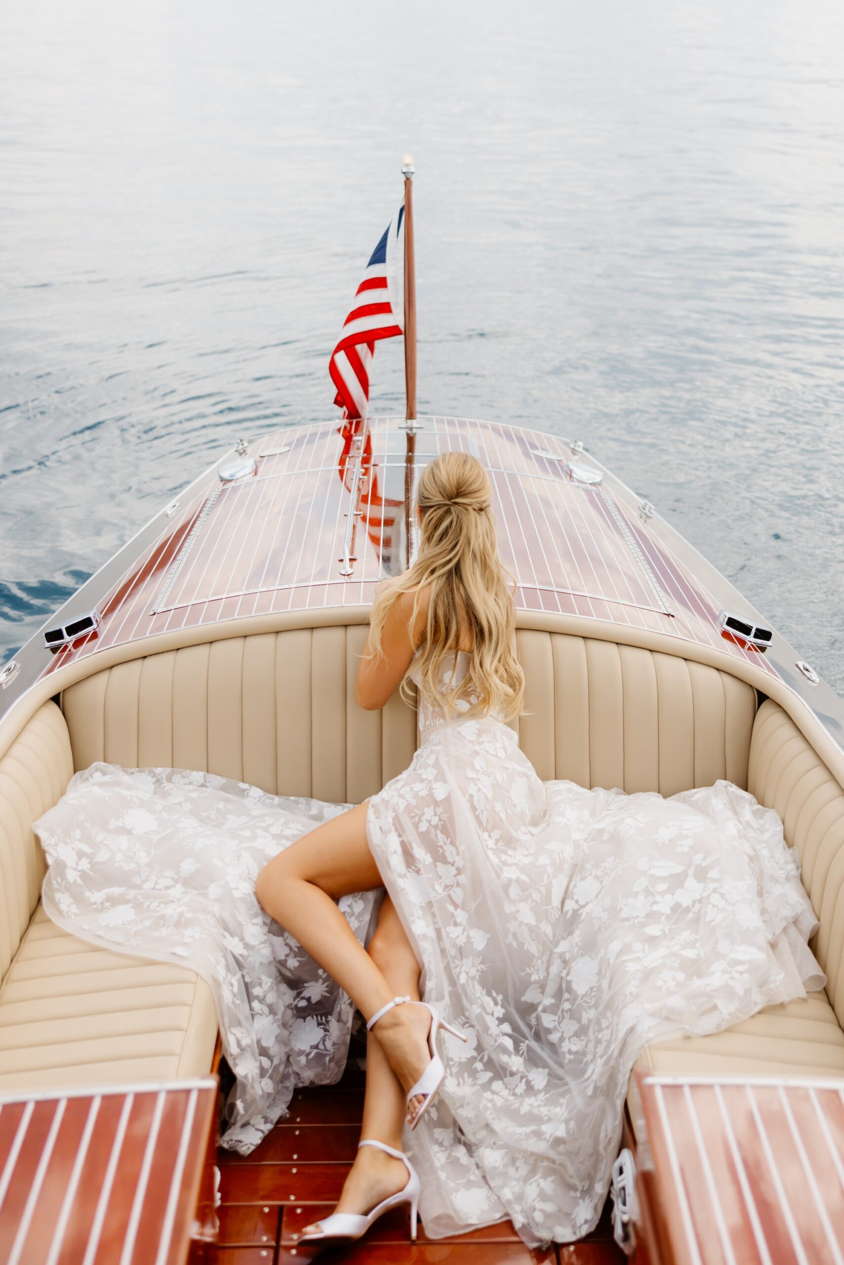 A woman in a white, flowing lace dress and heels sits on a wooden boat, facing