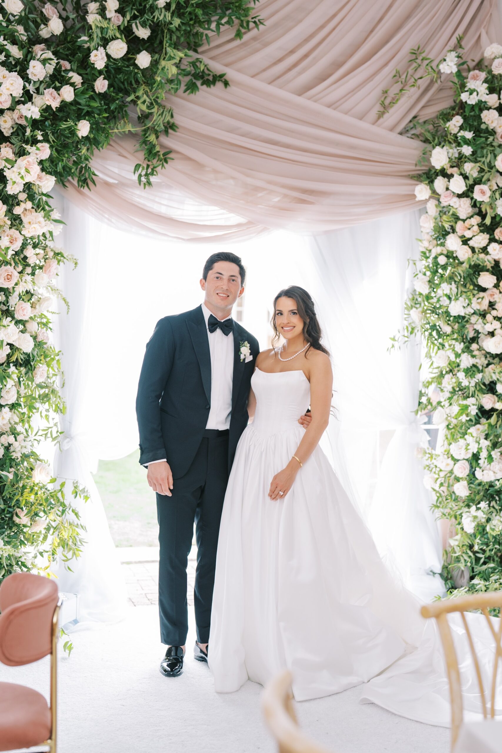 A bride in a white gown and a groom in a black tuxedo stand smiling A bride in a white gown and a groom in a black tuxedo stand smiling