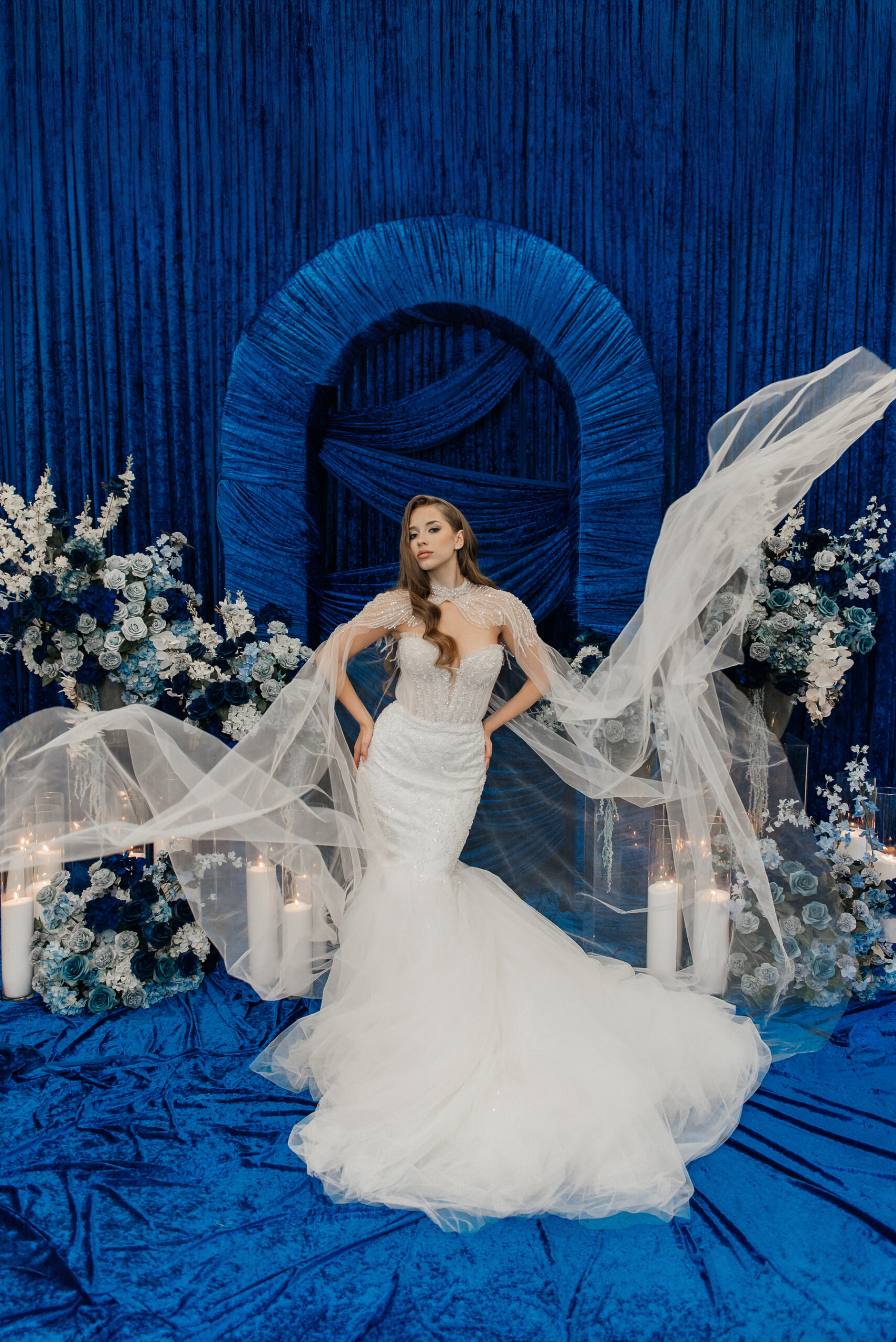 A bride in a white mermaid gown poses with a flowing veil against a deep Blue In Check backdrop, surrounded by blue and white flowers and tall candles. ~ WedLuxe