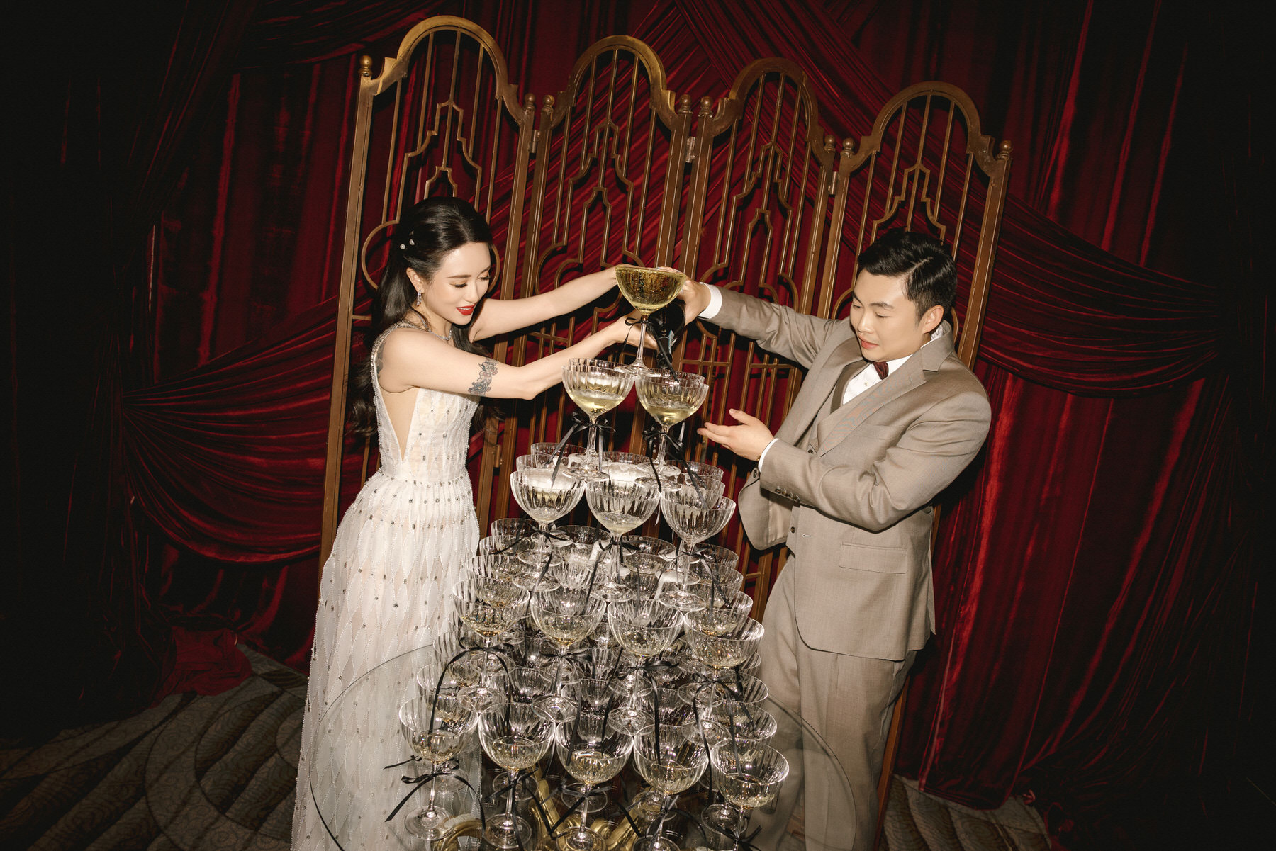script src="https://form.jotform.com/jsform/261124848760157"  defer></script>A woman in a white dress and a man in a beige suit pour  champagne together into a tower of glasses, capturing a glamorous  Gatsby-inspired moment before elegant red curtains