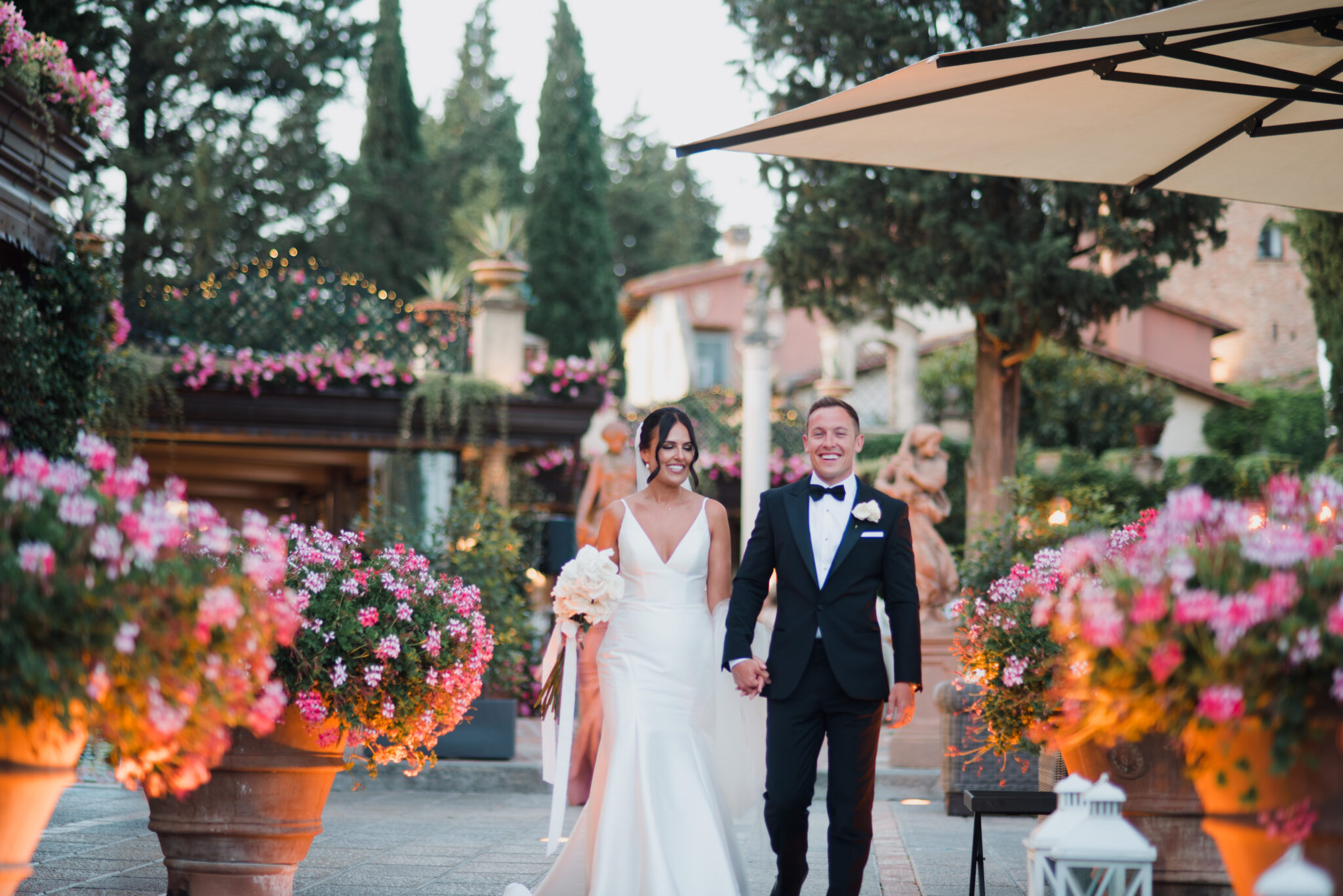 A bride and groom walk hand in hand outdoors, smiling. The elegant ...