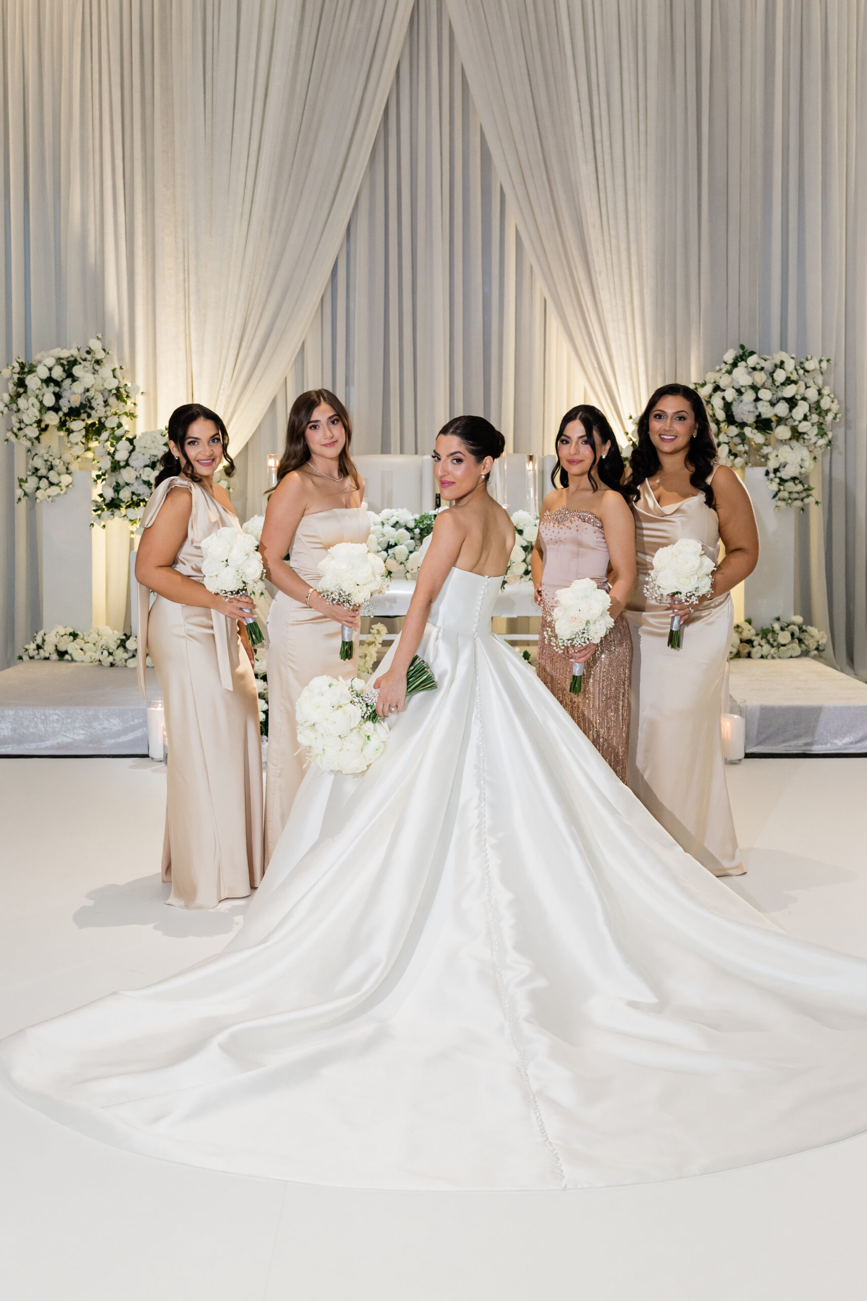 A bride in a white gown stands with four bridesmaids in light-colored dresses, all holding white bouquets, exuding elegance in an ornate ballroom wedding adorned with white flowers and draped curtains. ~ A bride in a white gown stands with four bridesmaids in light-colored dresses, all holding white bouquets, exuding elegance in an ornate ballroom wedding adorned with white flowers and draped curtains. ~