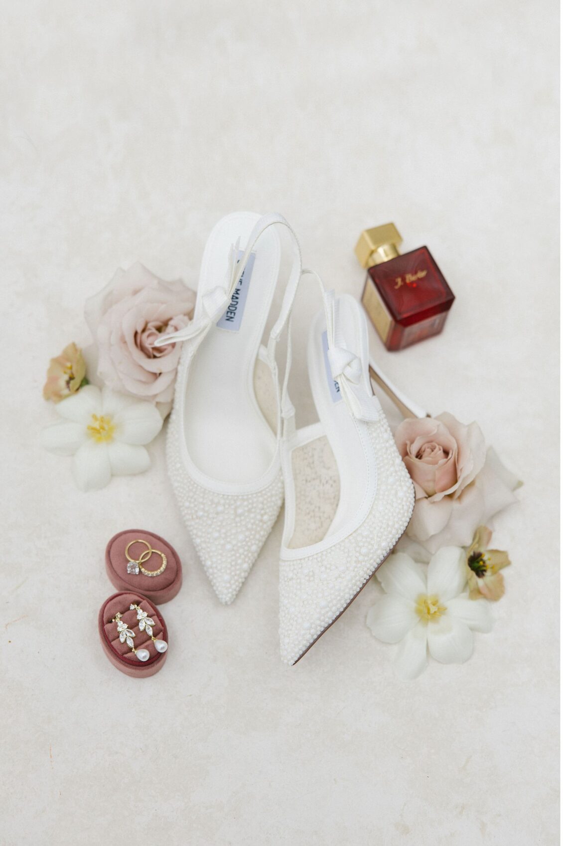 White bridal shoes, wedding rings, and perfume arranged with flowers