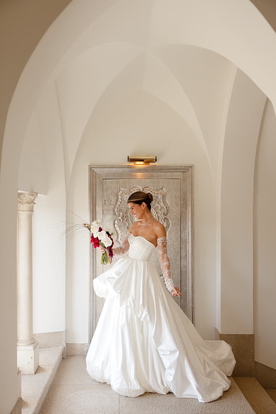 A bride in a white wedding dress stands near an ornate door, holding a bouquet of